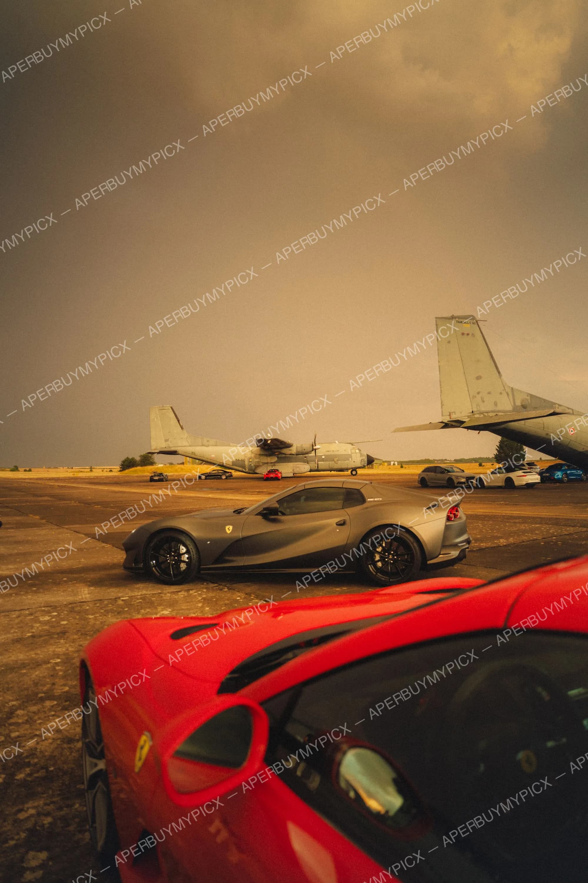 Ferrari 812 GTS grise mat garée sur un tarmac d'aérodrome militaire avec deux avions Transall C-160 en arrière-plan sous un ciel orageux, avant-plan d'une Ferrari rouge