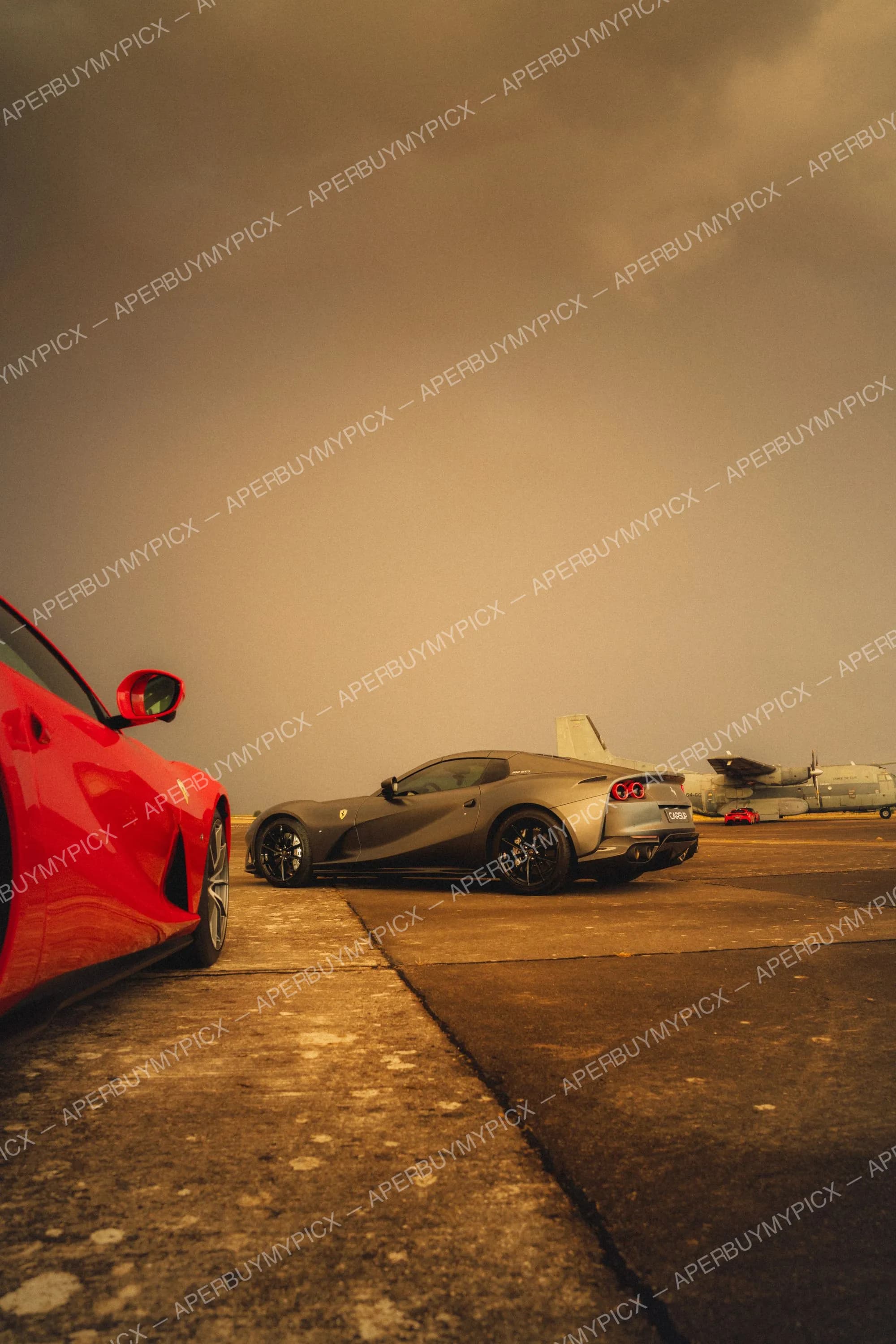 Ferrari 812 GTS grise vue de trois quarts arrière garée sur une piste d'aéroport militaire, à côté d'une Ferrari rouge au premier plan, avec des avions de transport et un ciel nuageux au crépuscule.