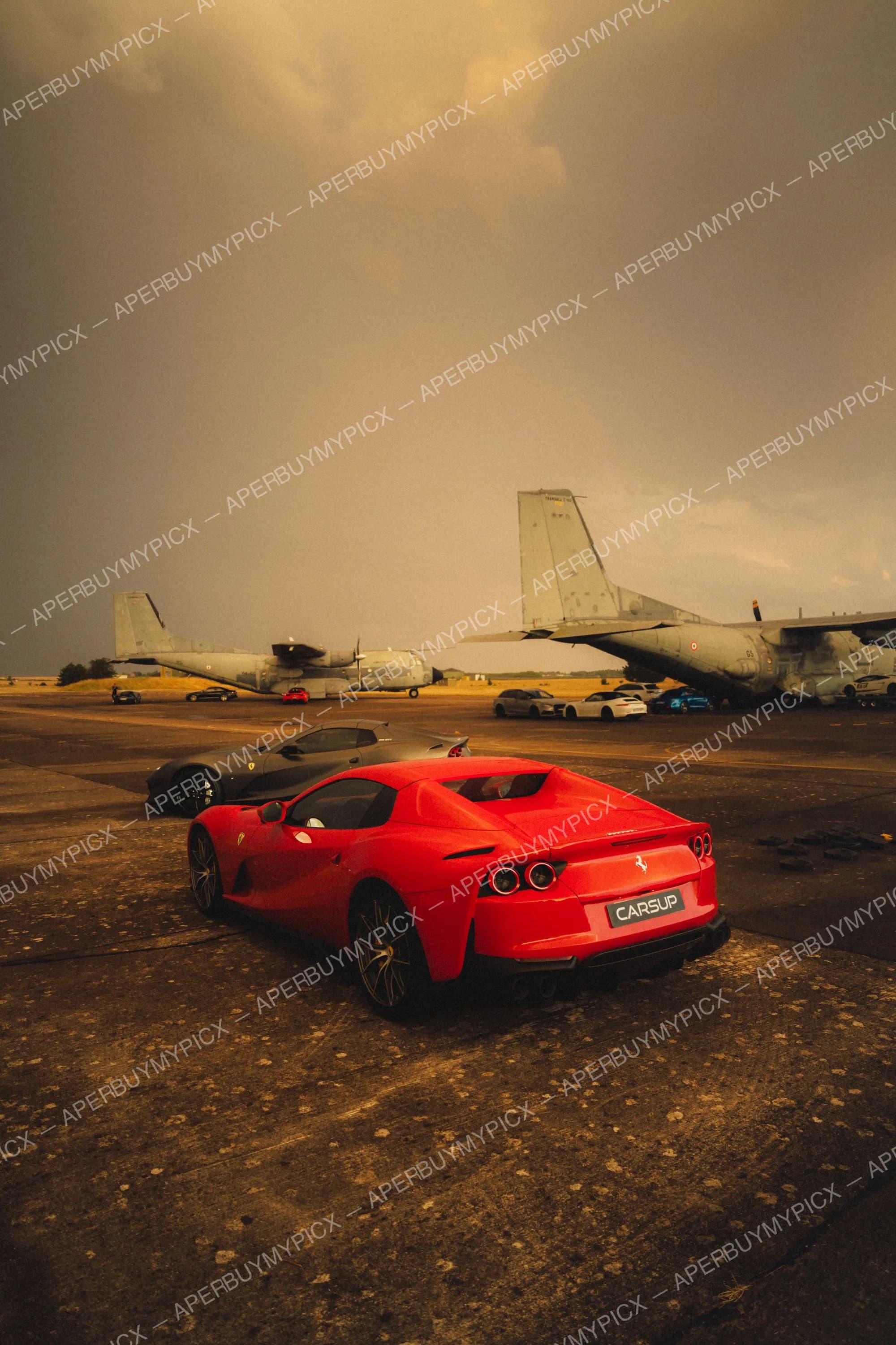Ferrari 812 rouge vue arrière garée sur un tarmac de base aérienne avec une Ferrari noire et des avions de transport militaire sous un ciel orageux au coucher du soleil