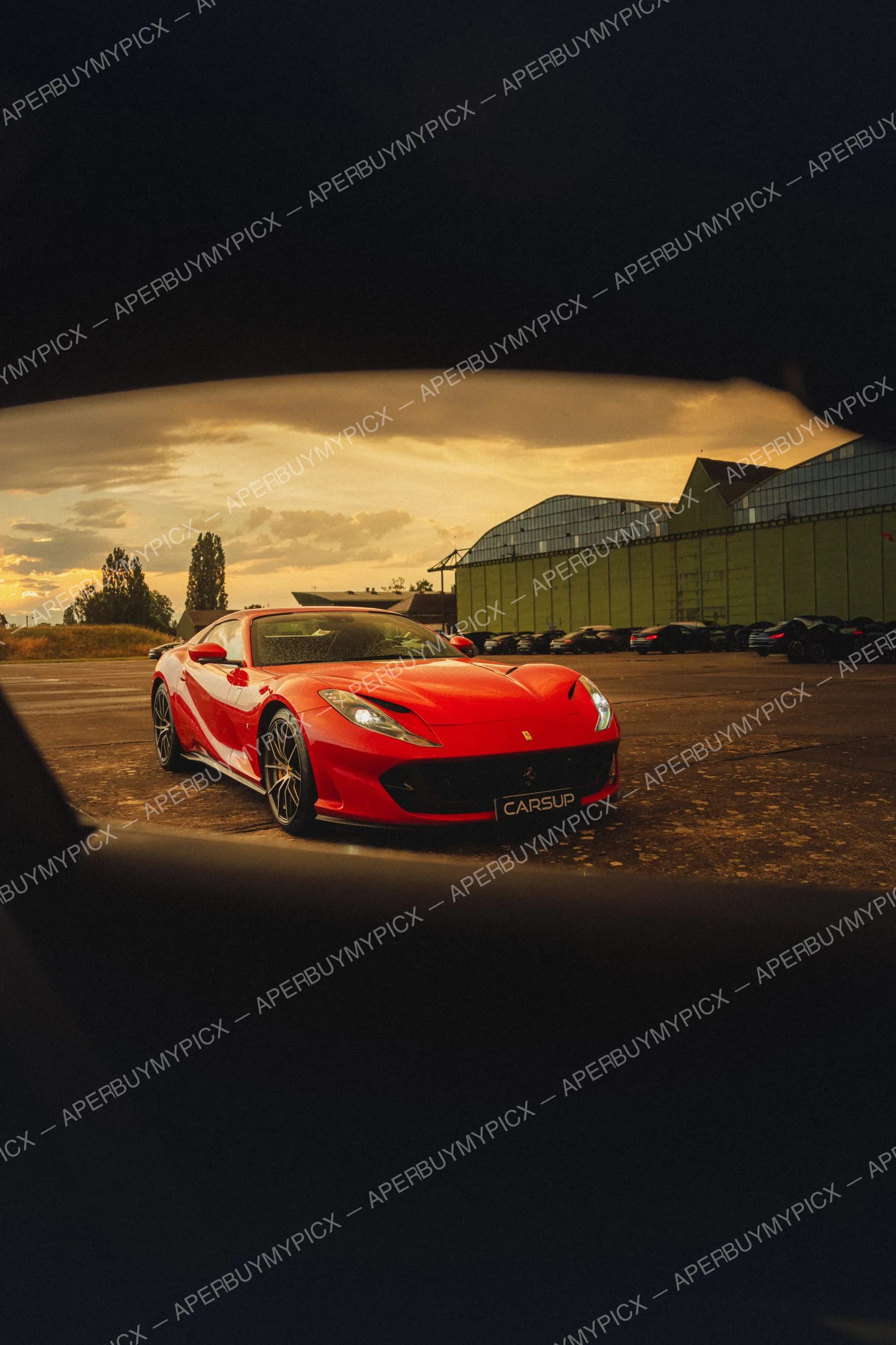Ferrari 812 Superfast rouge stationnée sur un tarmac devant un hangar vert au coucher de soleil, photographiée à travers l'ouverture d'une fenêtre