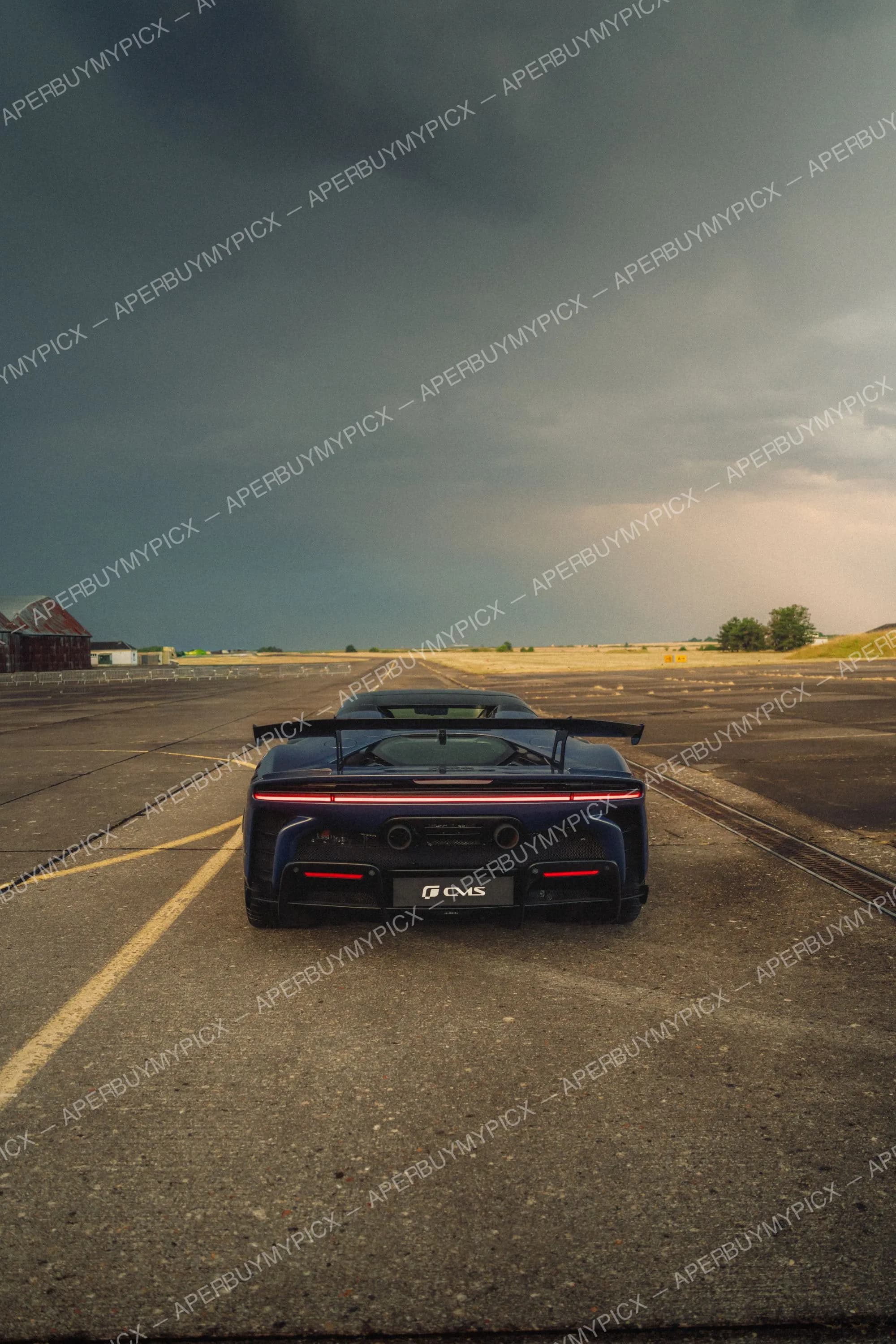 Vue arrière d'une supercar bleu foncé avec aileron et feux LED, stationnée sur une piste d'aérodrome sous un ciel sombre et orageux.