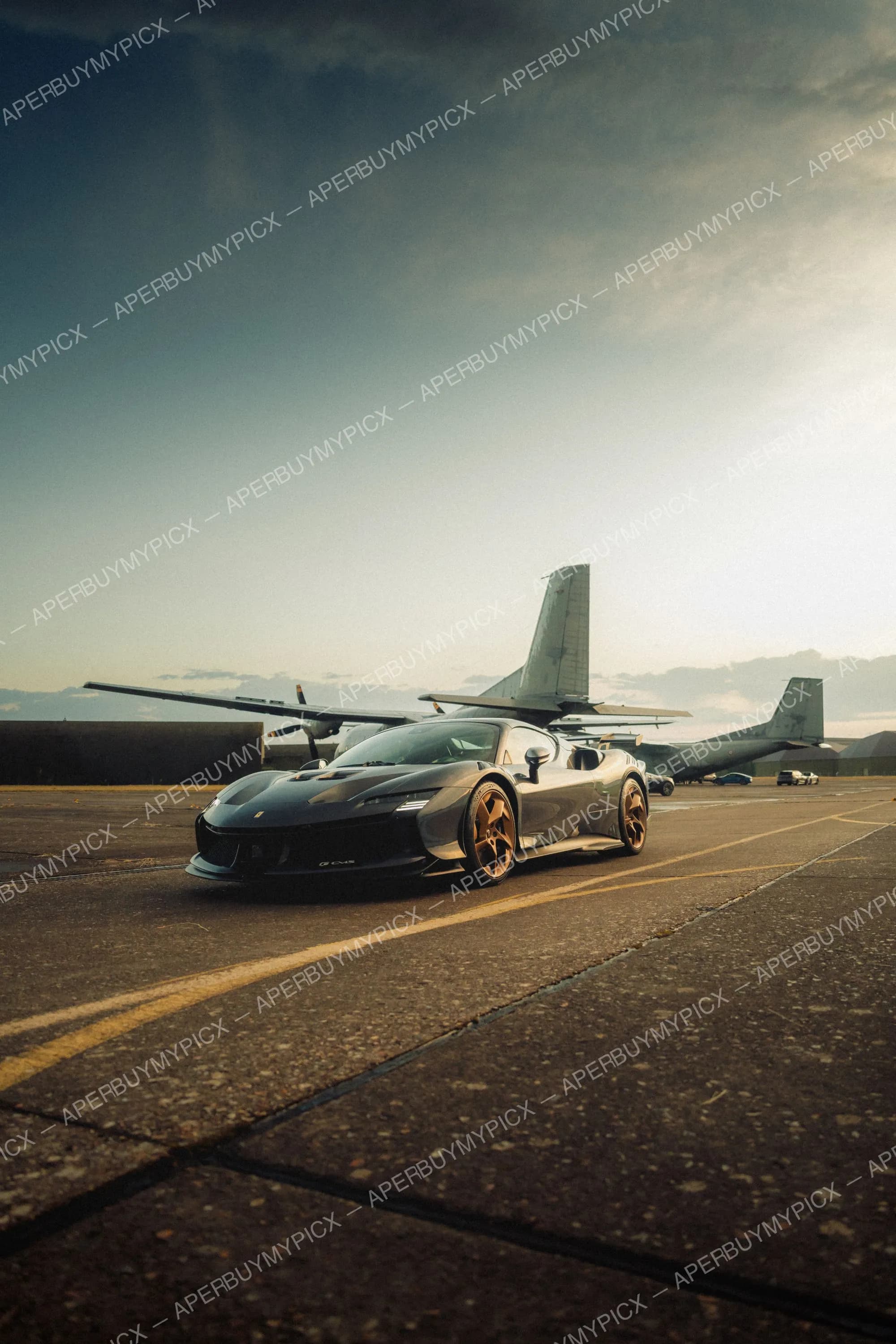 Ferrari SF90 XX grise aux jantes bronze garée sur une piste d'aviation, devant des avions militaires à hélices, sous un ciel nuageux au coucher du soleil