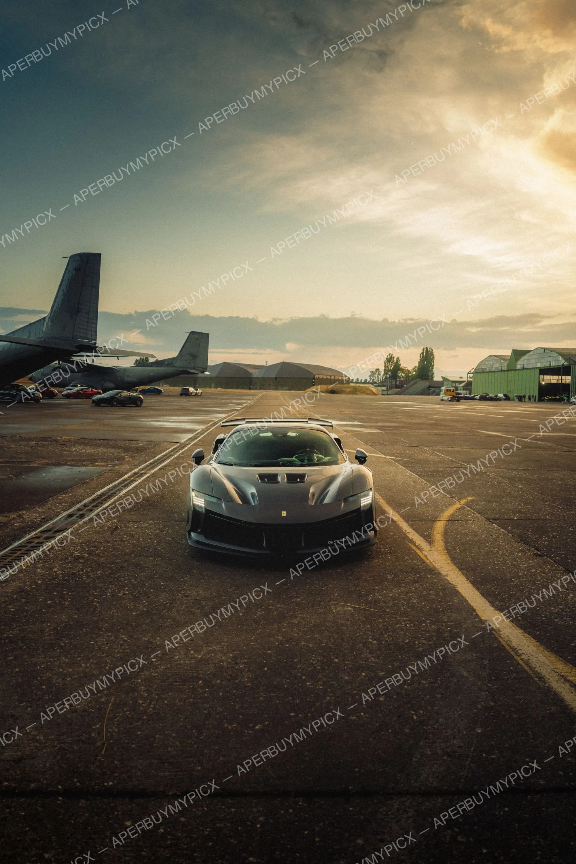 Ferrari SF90 XX grise vue de face sur une piste d'aérodrome au coucher du soleil, avec des avions militaires et des hangars en arrière-plan sous un ciel nuageux doré.