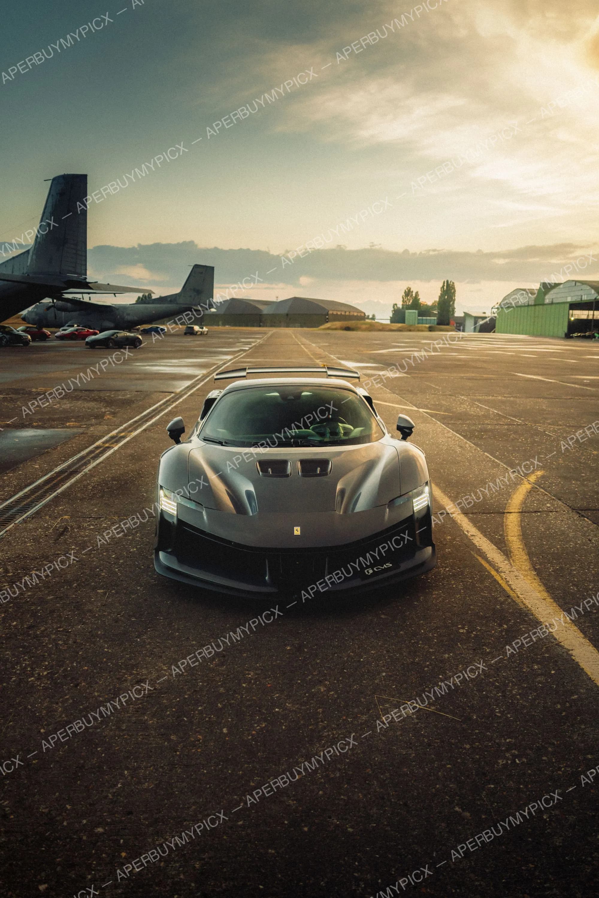 Ferrari SF90 grise vue de face sur une piste d'aéroport avec avions militaires et hangars au coucher du soleil