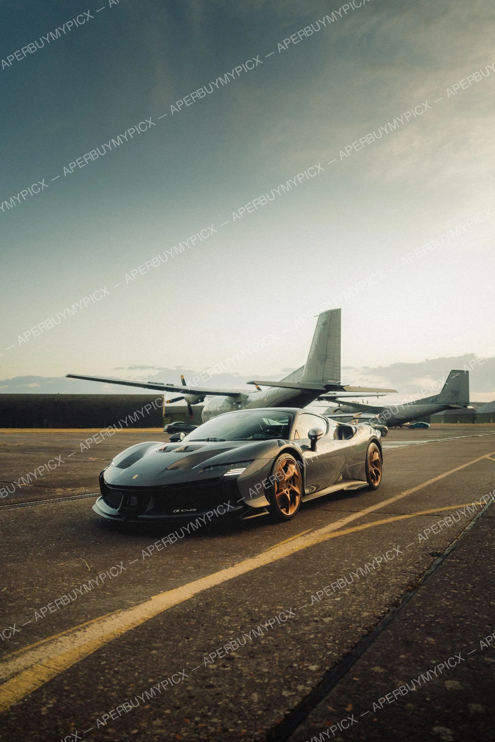 Ferrari SF90 XX grise aux jantes bronze garée sur une piste d'aéroport militaire devant des avions cargo à hélices au coucher du soleil