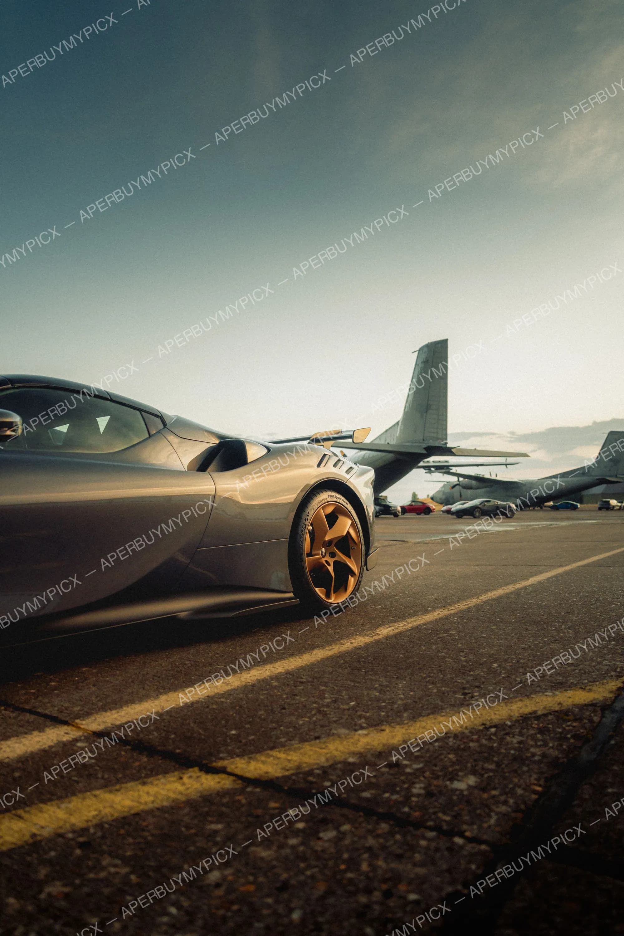 Supercar grise avec jantes dorées vue de profil sur le tarmac d'un aéroport, avec des avions militaires et voitures en arrière-plan au coucher du soleil