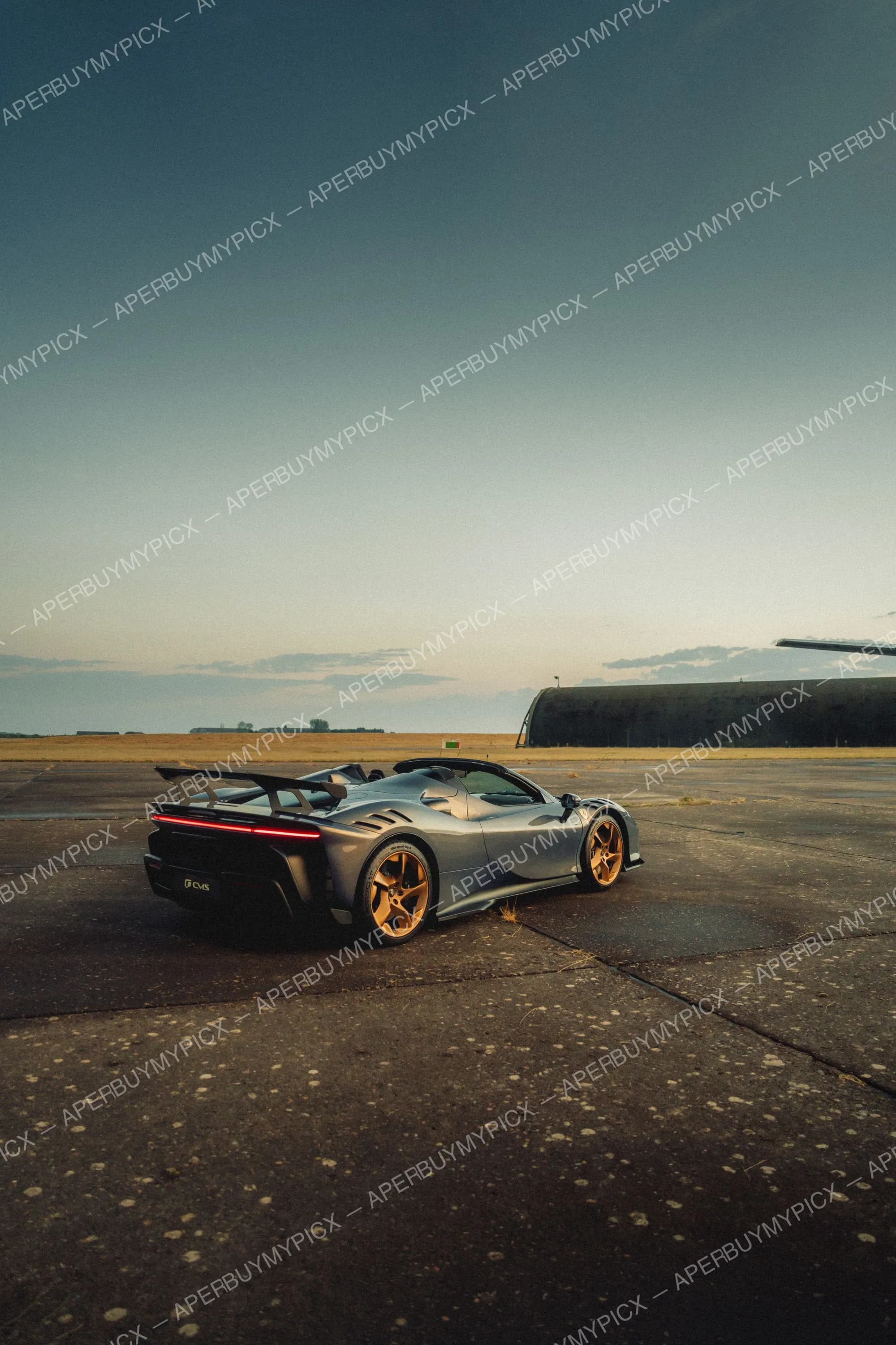Ferrari SF90 Spider grise aux jantes dorées garée sur une piste d'aérodrome au coucher du soleil, avec un hangar en arrière-plan