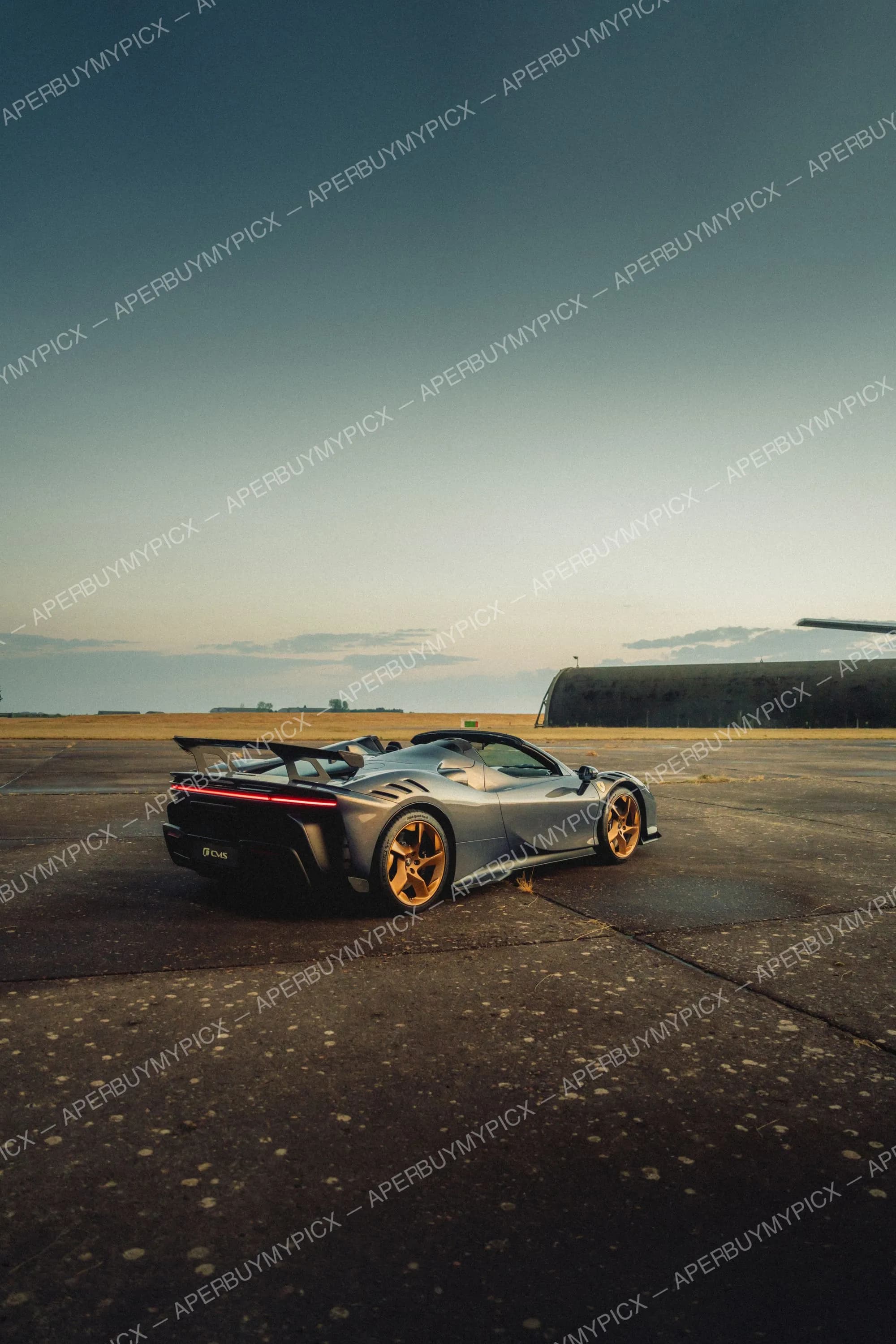 Ferrari SF90 Spider grise avec jantes dorées et aileron arrière, garée sur une piste d'aérodrome avec hangar au coucher du soleil