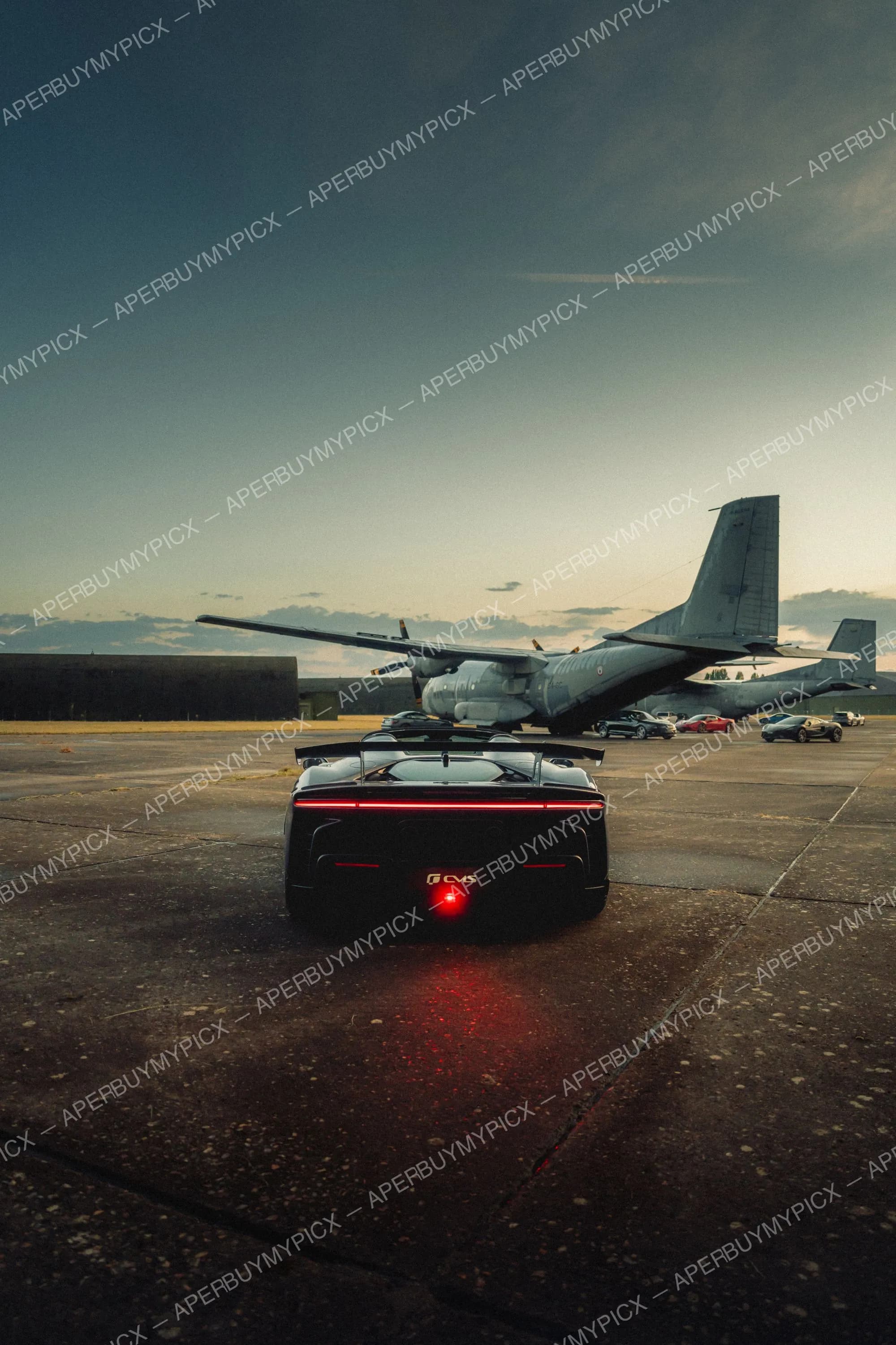 Vue arrière d'une supercar noire aux feux arrière rouges lumineux, garée sur un tarmac d'aérodrome face à un avion de transport militaire, au crépuscule, avec d'autres voitures de sport au loin.