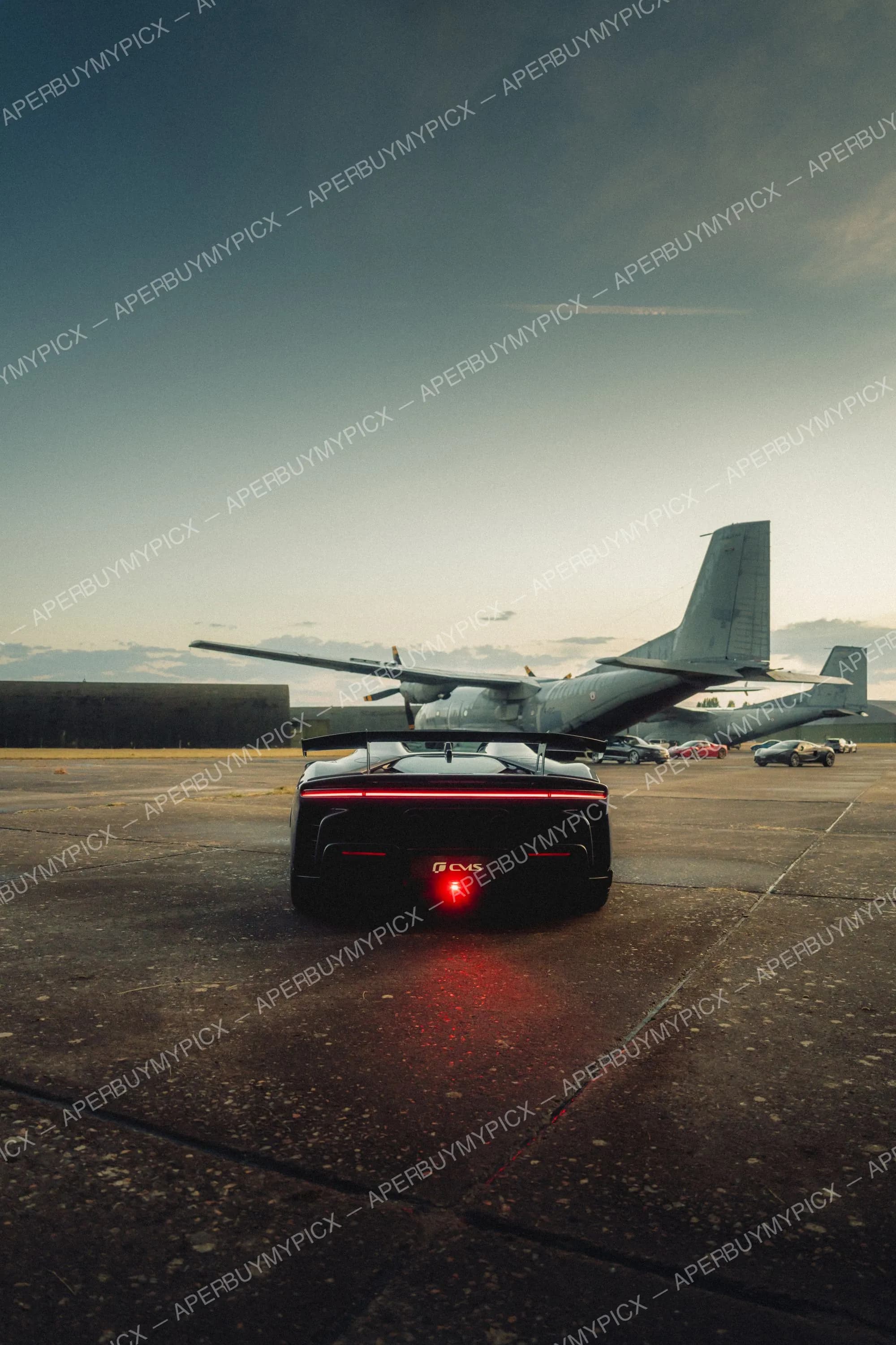 Vue arrière d'une supercar noire aux feux LED rouges garée sur un tarmac au coucher du soleil, face à un avion de transport militaire et d'autres voitures de sport.