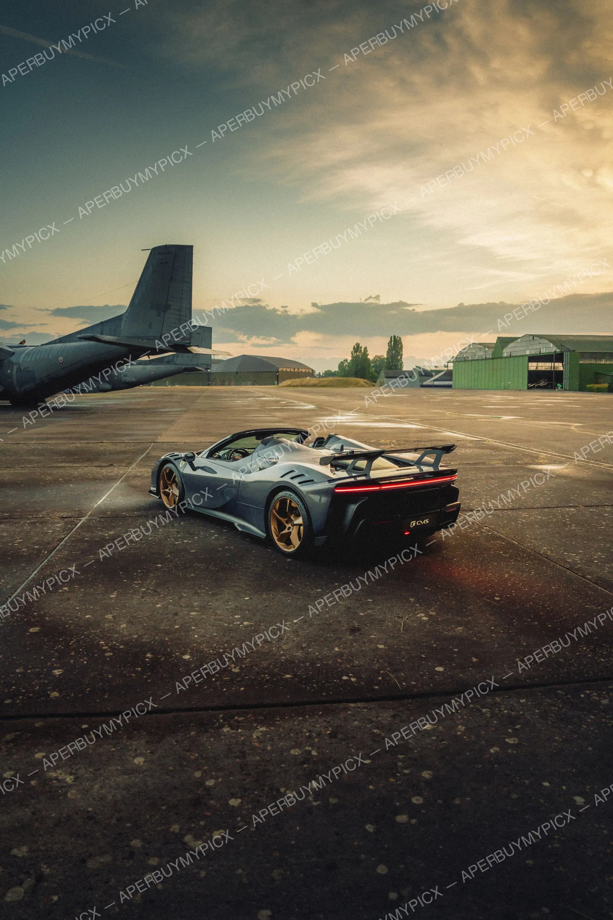 Ferrari SF90 Spider grise aux jantes dorées vue de trois quarts arrière sur un tarmac de base aérienne, avec un avion militaire et des hangars verts au coucher du soleil