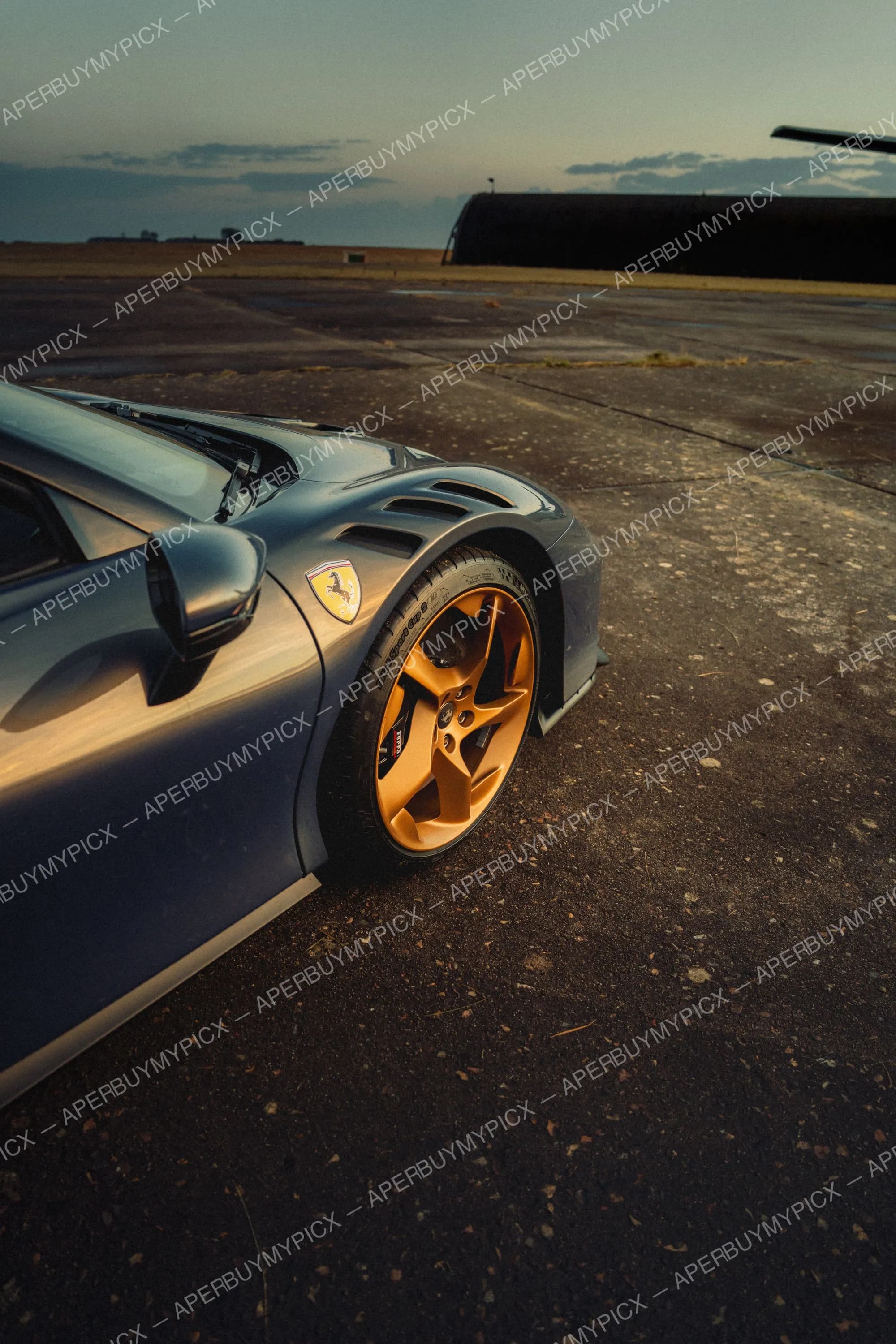 Gros plan sur l'aile avant et la jante dorée d'une Ferrari grise avec écusson Cheval Cabré, garée sur une piste d'aérodrome au coucher du soleil avec un hangar en arrière-plan.