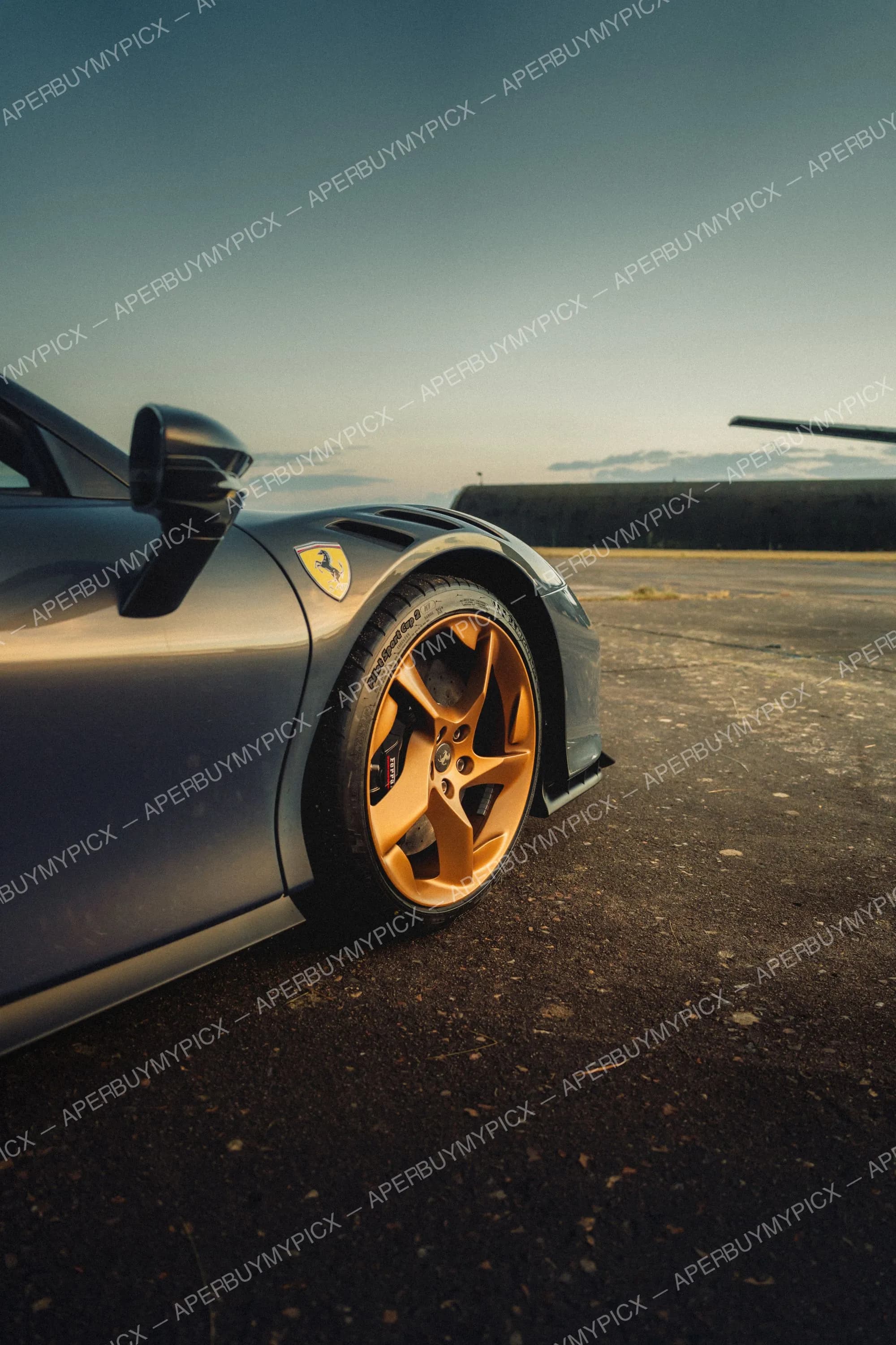 Gros plan sur la roue avant droite d'une Ferrari grise avec jante dorée et étrier de frein rouge, garée sur une piste bétonnée au coucher du soleil avec un hangar en arrière-plan.