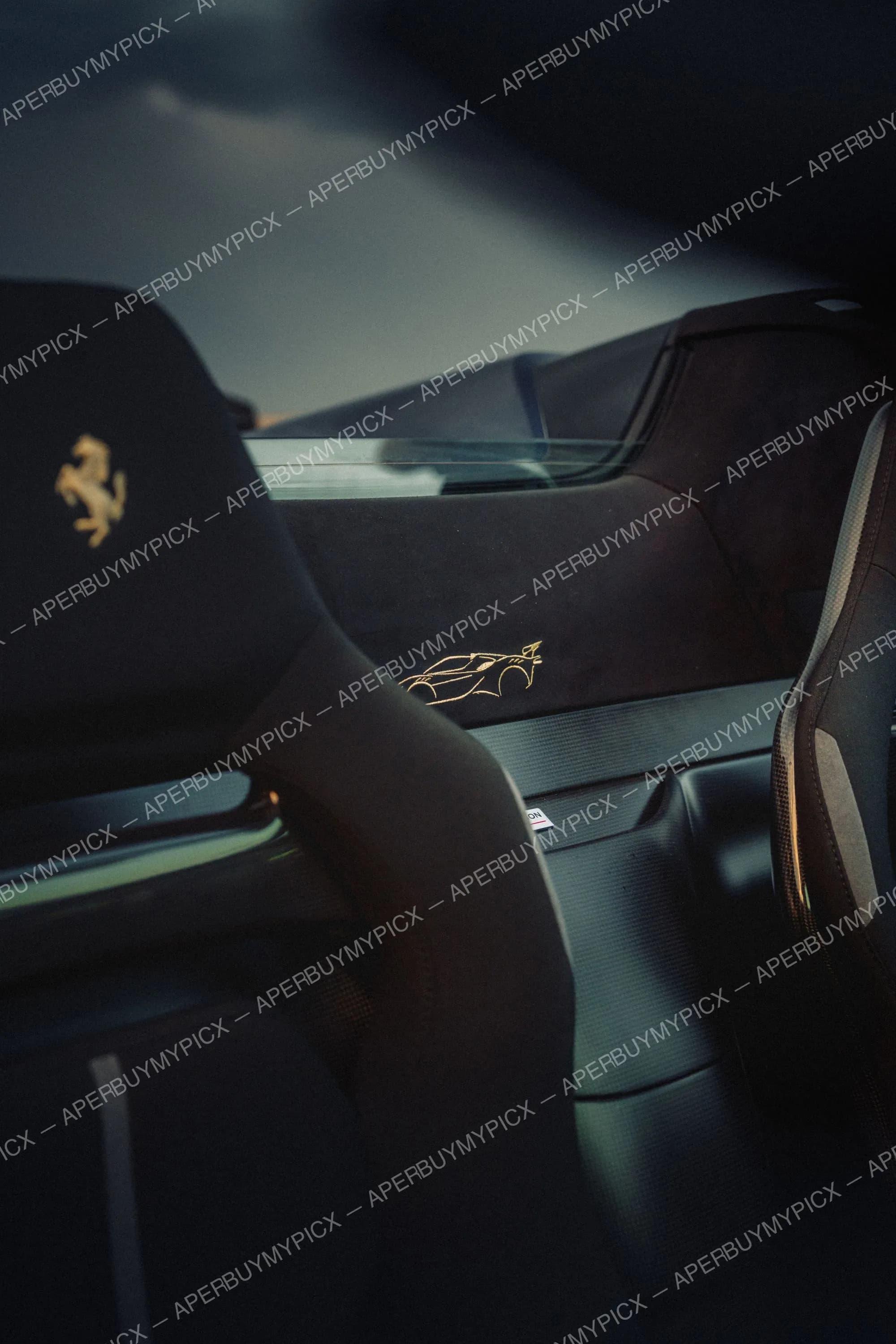 Gros plan sur les sièges baquets en Alcantara noir d'une Ferrari, avec le logo du cheval cabré brodé en doré sur l'appui-tête et une silhouette de voiture de course brodée en or, dans une lumière tamisée.