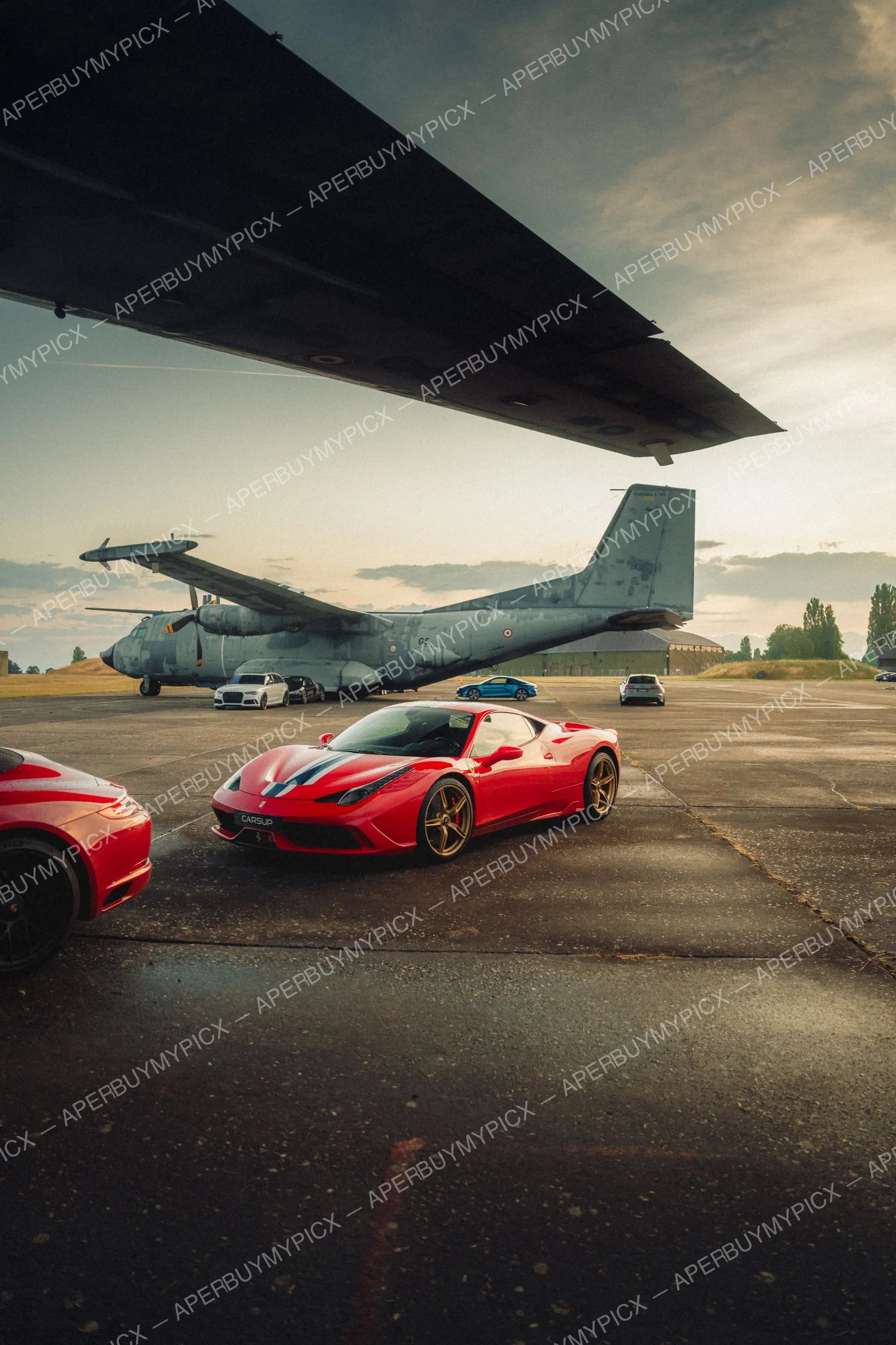 Ferrari 458 Speciale rouge à bandes tricolores stationnée sur le tarmac sous l'aile d'un avion militaire Transall C-160, avec d'autres voitures sportives et un ciel nuageux au coucher du soleil.