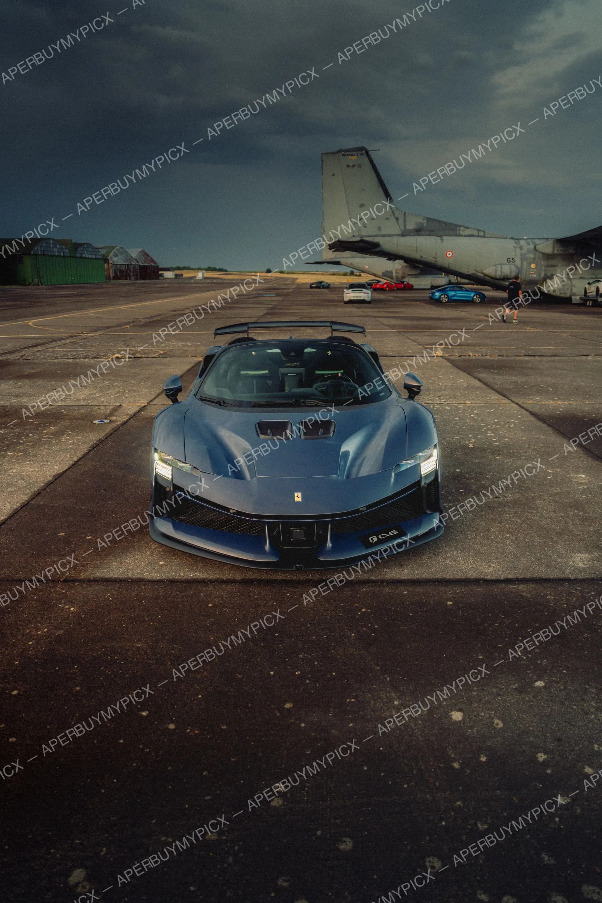 Ferrari SF90 XX bleue vue de face garée sur une piste d'aéroport avec aileron arrière, devant un avion militaire et d'autres voitures de sport sous un ciel orageux