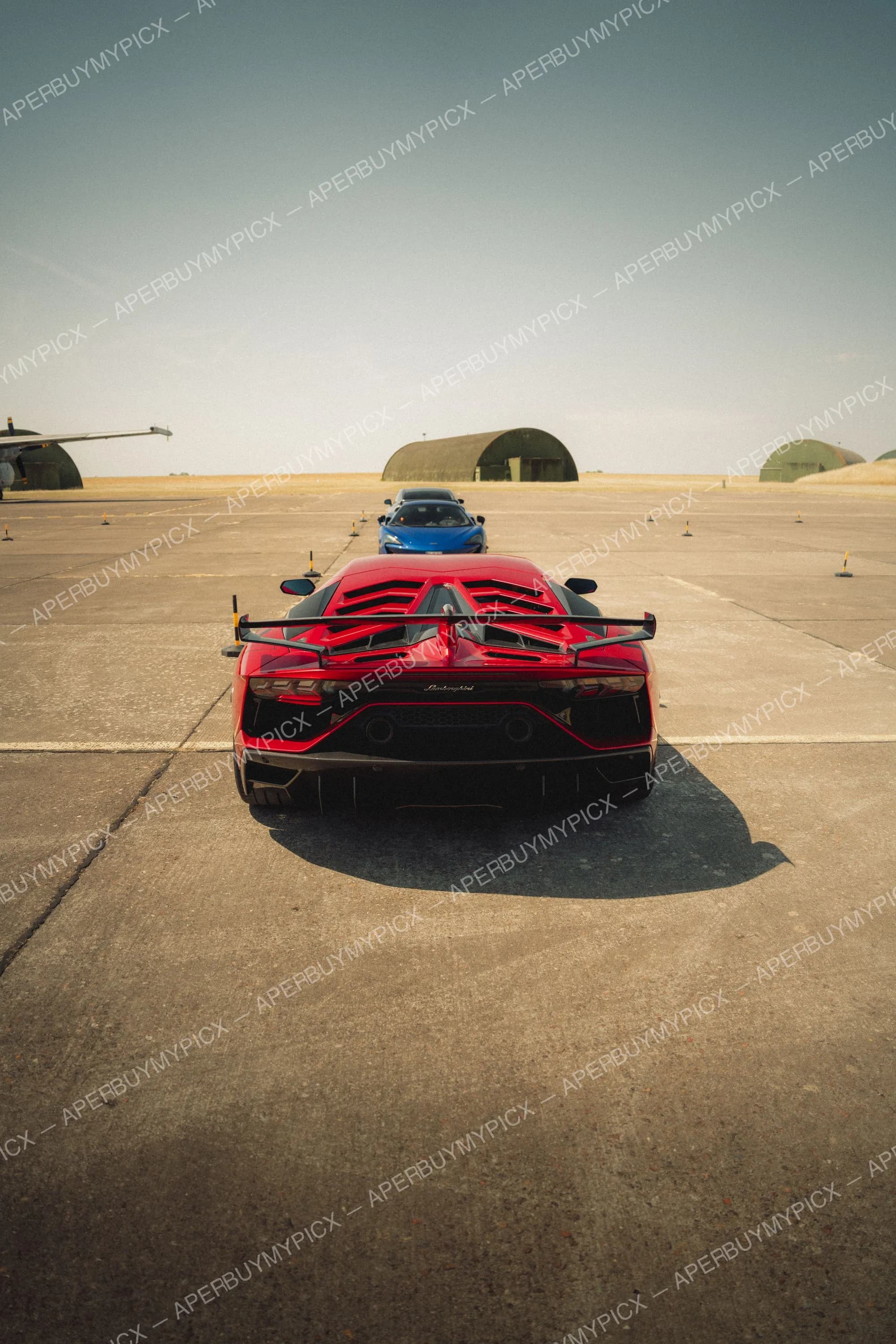 Vue arrière d'une Lamborghini Aventador SVJ rouge avec aileron noir, garée sur une piste en béton, suivie d'une McLaren bleue et d'une voiture grise, avec hangars militaires et ciel dégagé en arrière-plan.