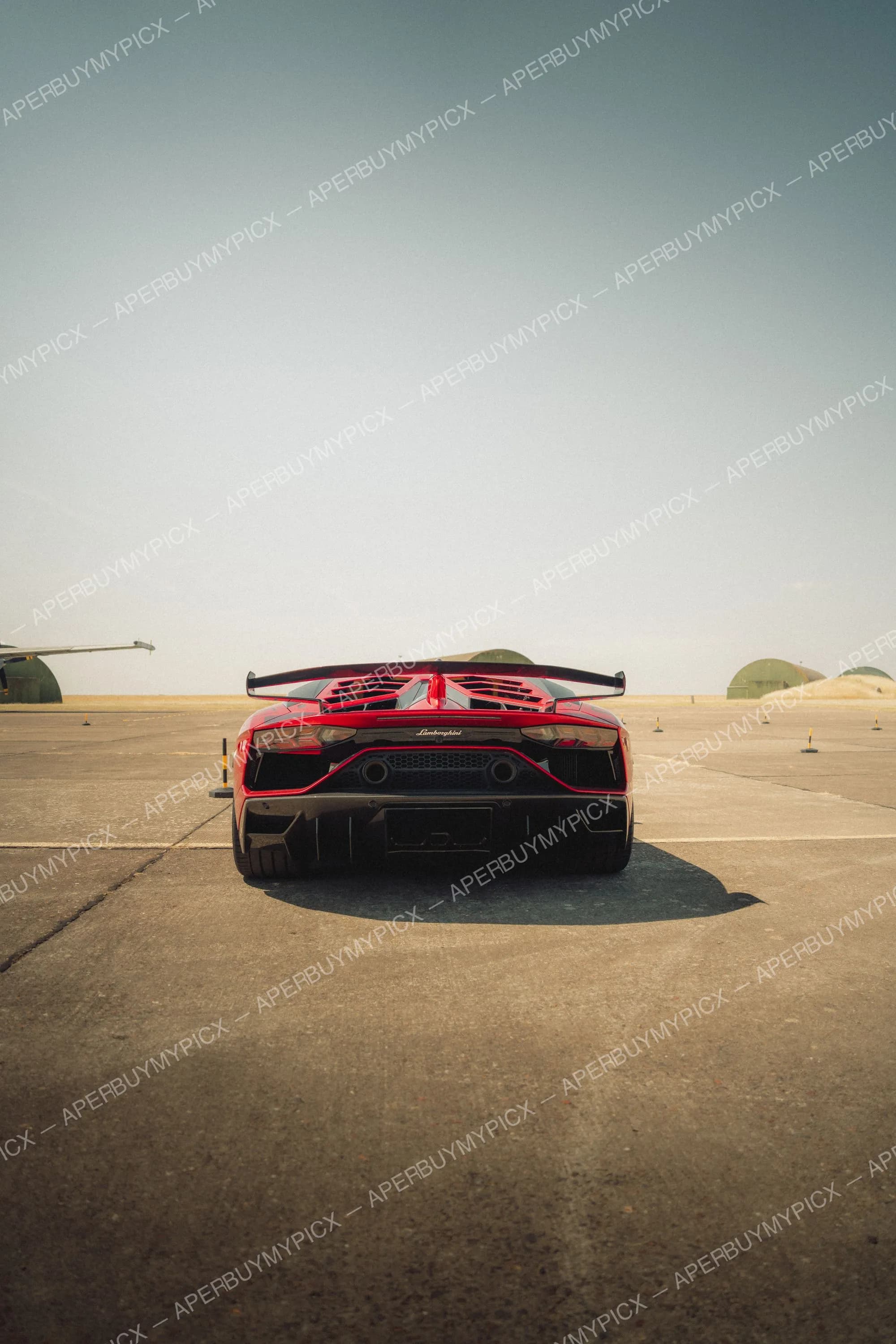 Lamborghini Aventador SVJ rouge vue de l'arrière avec aileron sur une piste d'aérodrome, hangars et ciel clair en arrière-plan