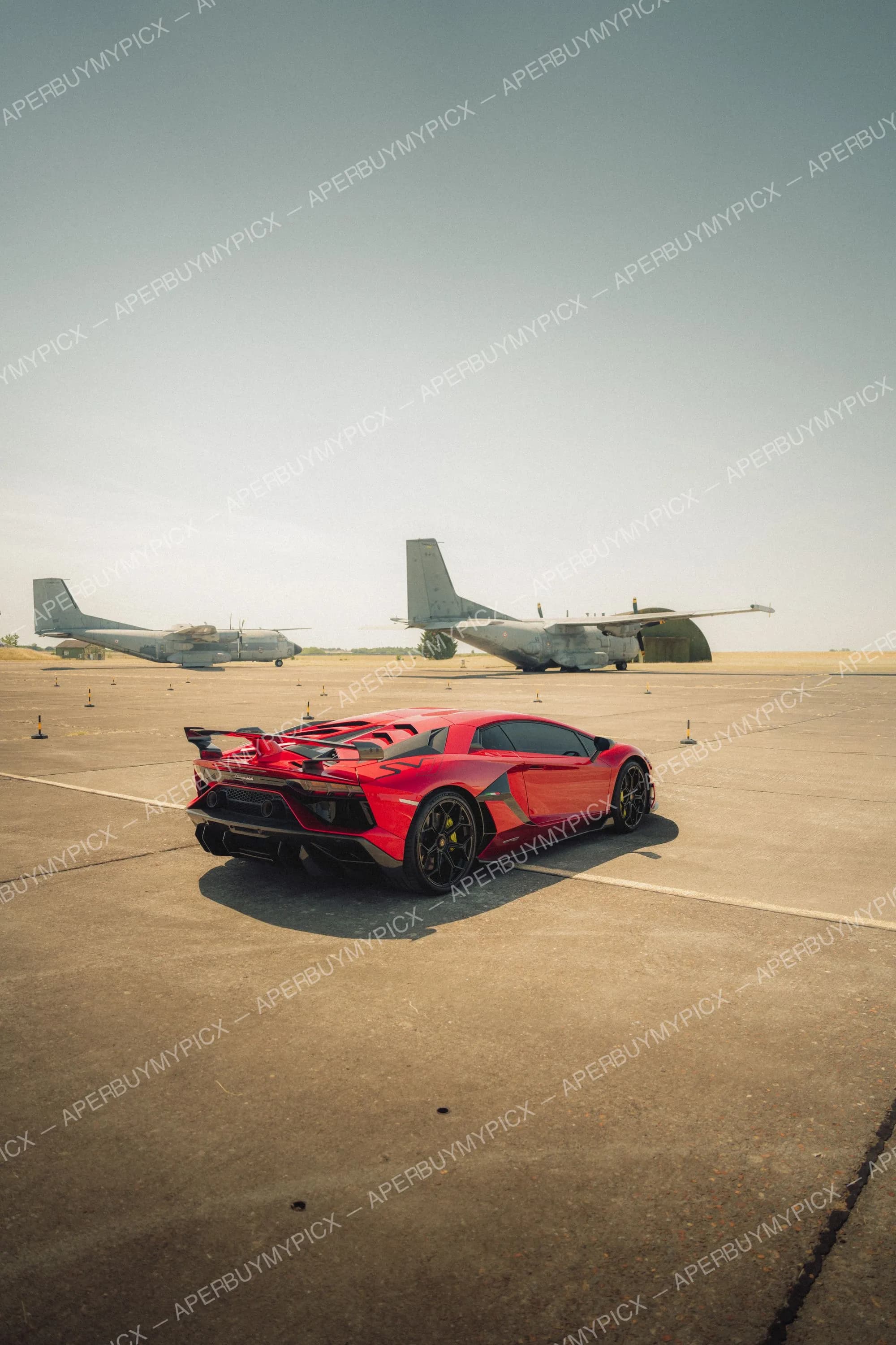 Lamborghini Aventador SVJ rouge vue arrière sur le tarmac d'une base aérienne, deux avions de transport militaire en arrière-plan sous un ciel dégagé