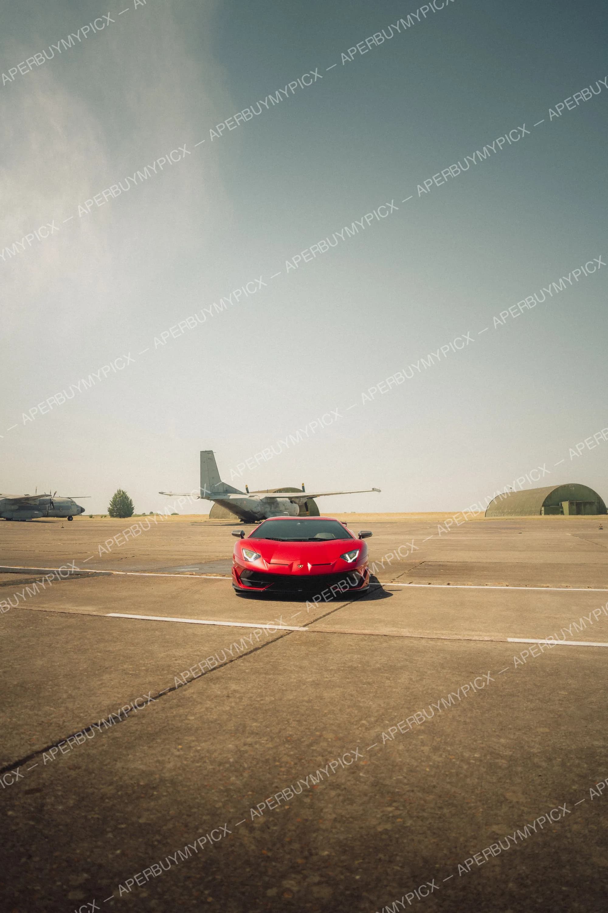 Lamborghini Aventador SVJ rouge stationnée sur le tarmac d'une base aérienne, avec des avions militaires et un hangar en arrière-plan sous un ciel voilé.
