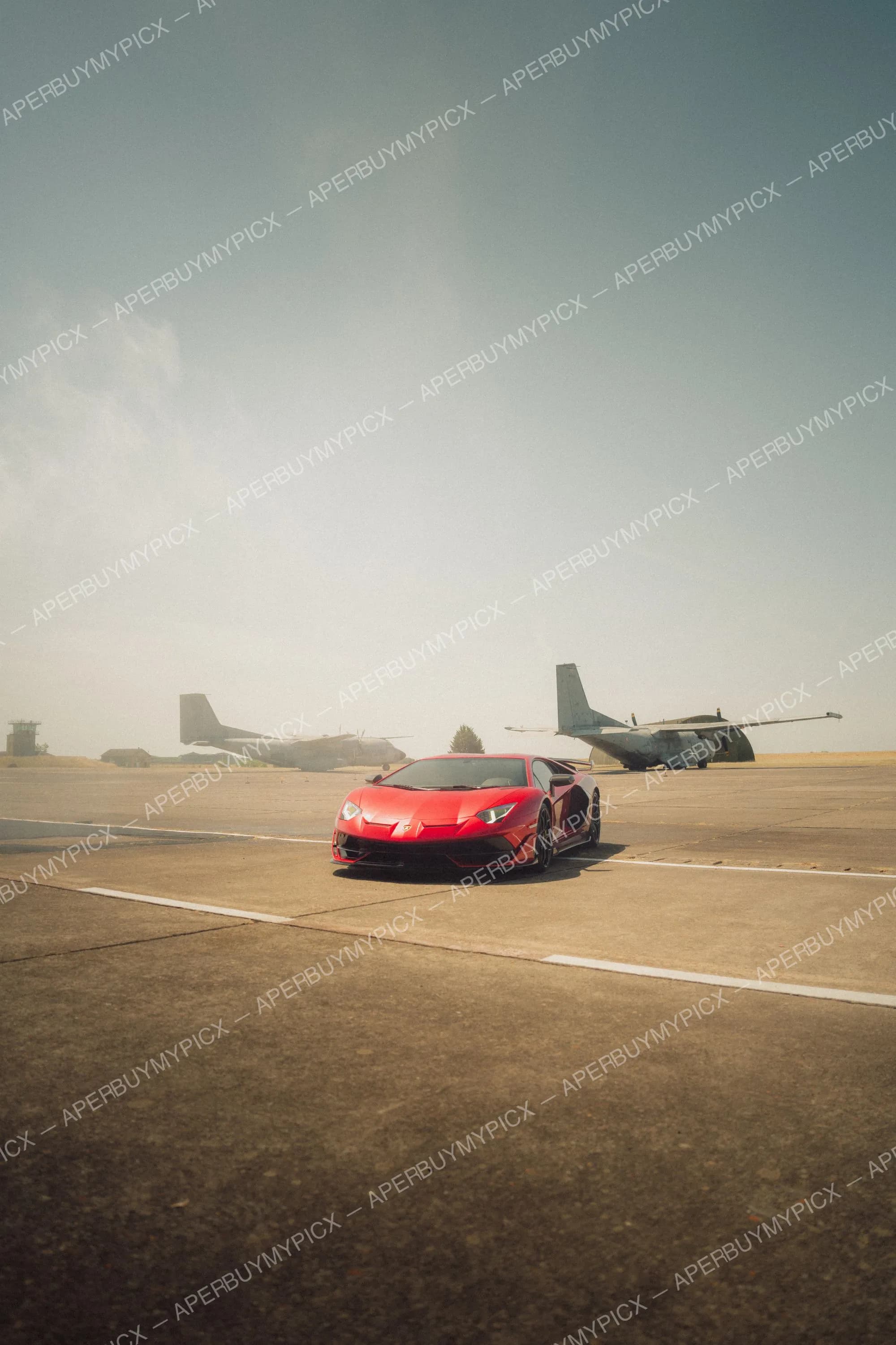 Lamborghini Aventador SVJ rouge stationnée sur une piste d'aérodrome avec deux avions de transport militaire à l'arrière-plan sous un ciel voilé