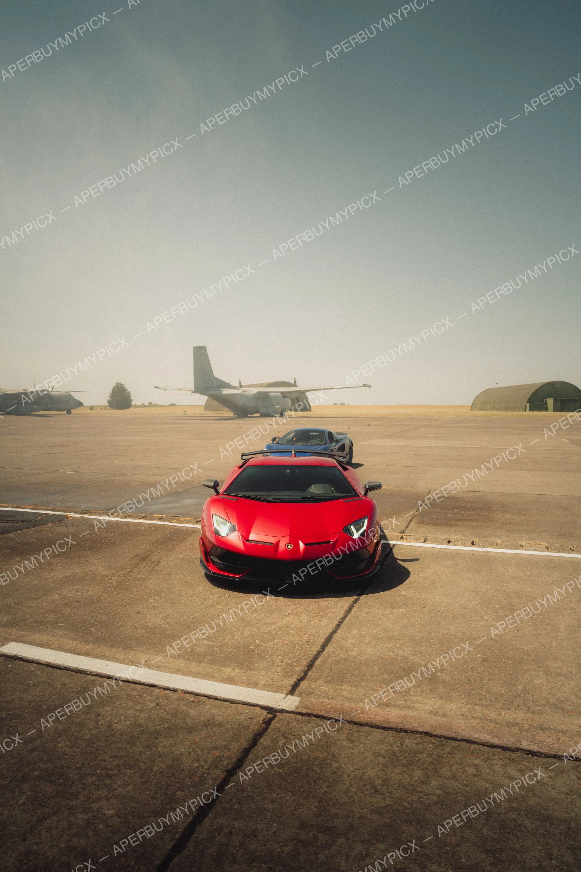Lamborghini Aventador SVJ rouge garée sur un tarmac d'aérodrome avec un avion militaire, un hangar et une supercar bleue en arrière-plan sous un ciel dégagé