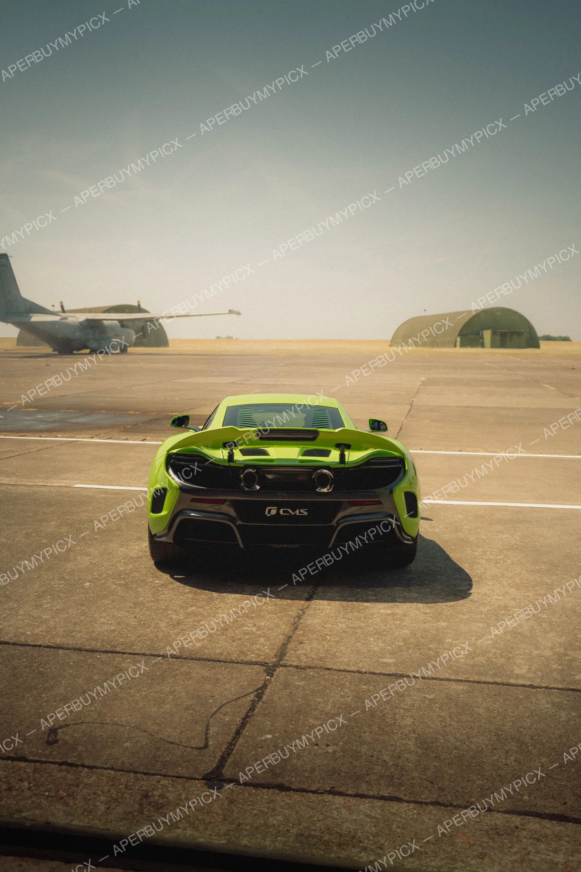 Vue arrière d'une supercar verte au diffuseur carbone et double échappement, garée sur le tarmac d'un aérodrome militaire avec un avion cargo et un hangar au loin sous un ciel dégagé.