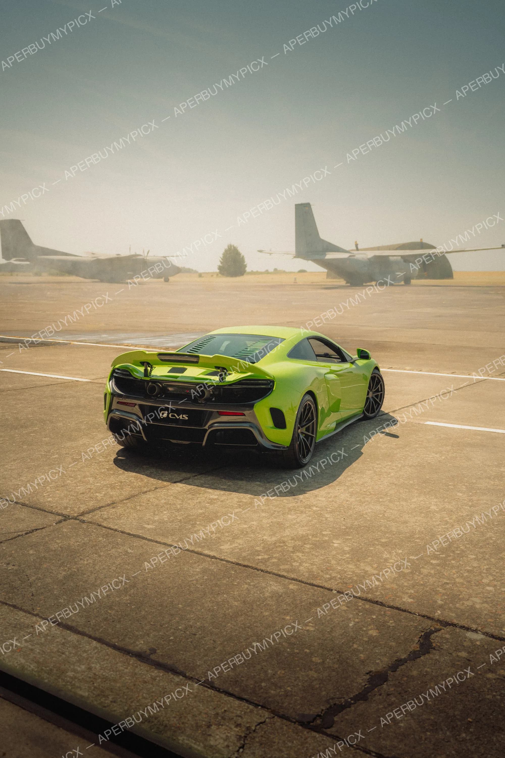McLaren 675LT vert vif vue arrière stationnée sur une piste d'aéroport avec deux avions de transport militaire en arrière-plan sous un ciel clair
