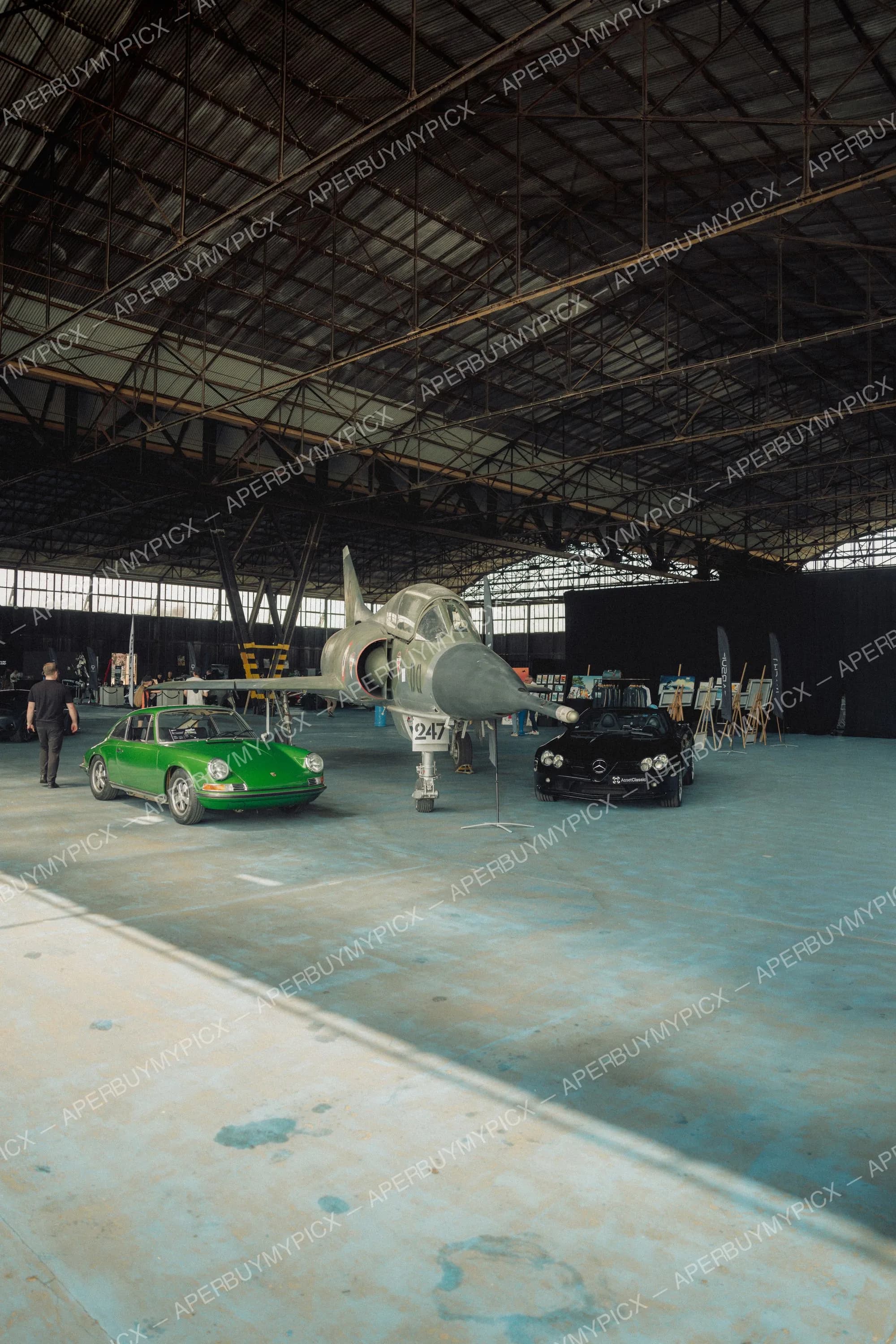 Porsche 911 verte classique et Mercedes noire garées à côté d'un chasseur Mirage gris dans un vaste hangar industriel avec expositions et visiteurs.