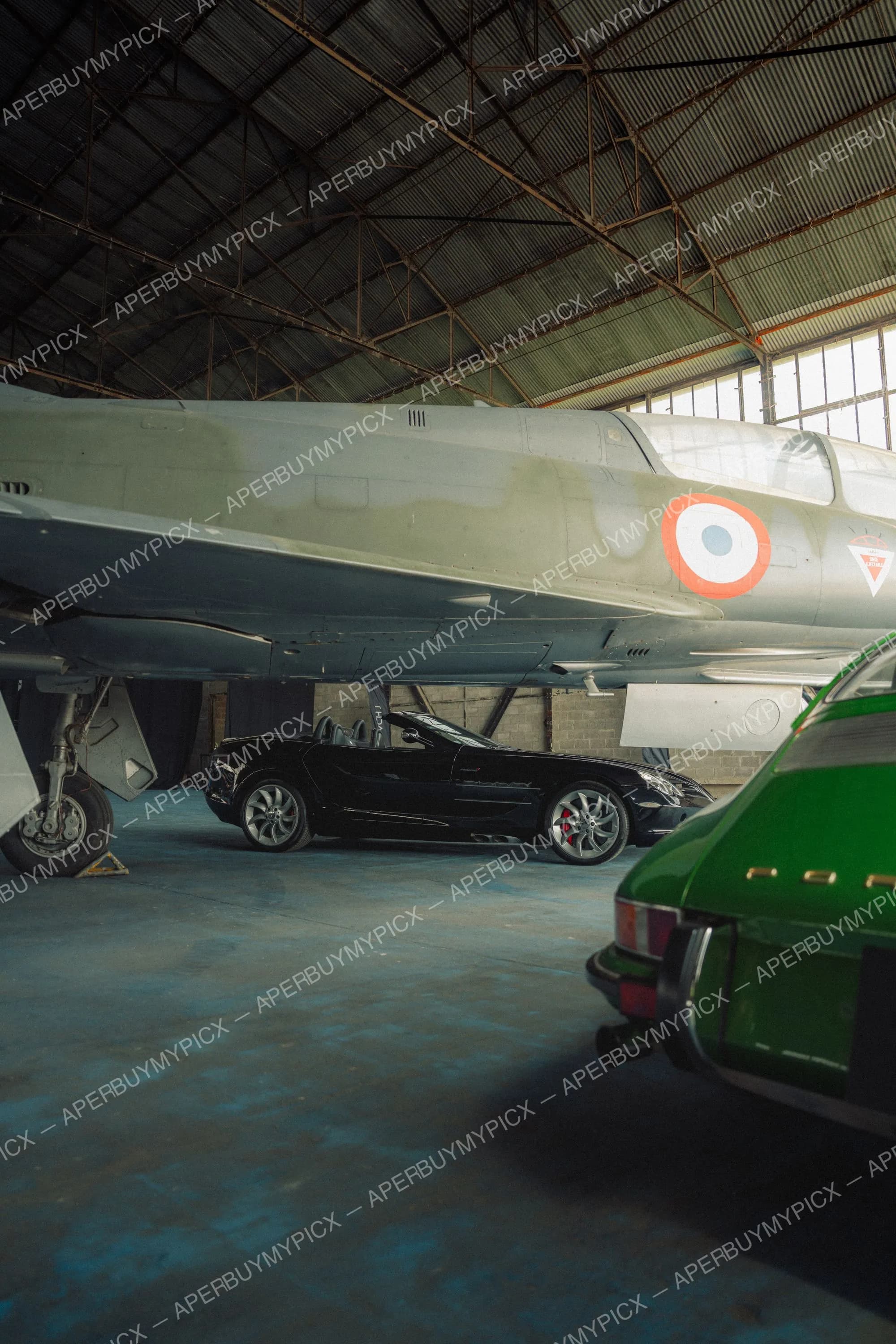 Une Mercedes SLR McLaren noire et une Porsche 911 verte garées dans un hangar d'aviation sous un avion de chasse militaire avec cocarde tricolore