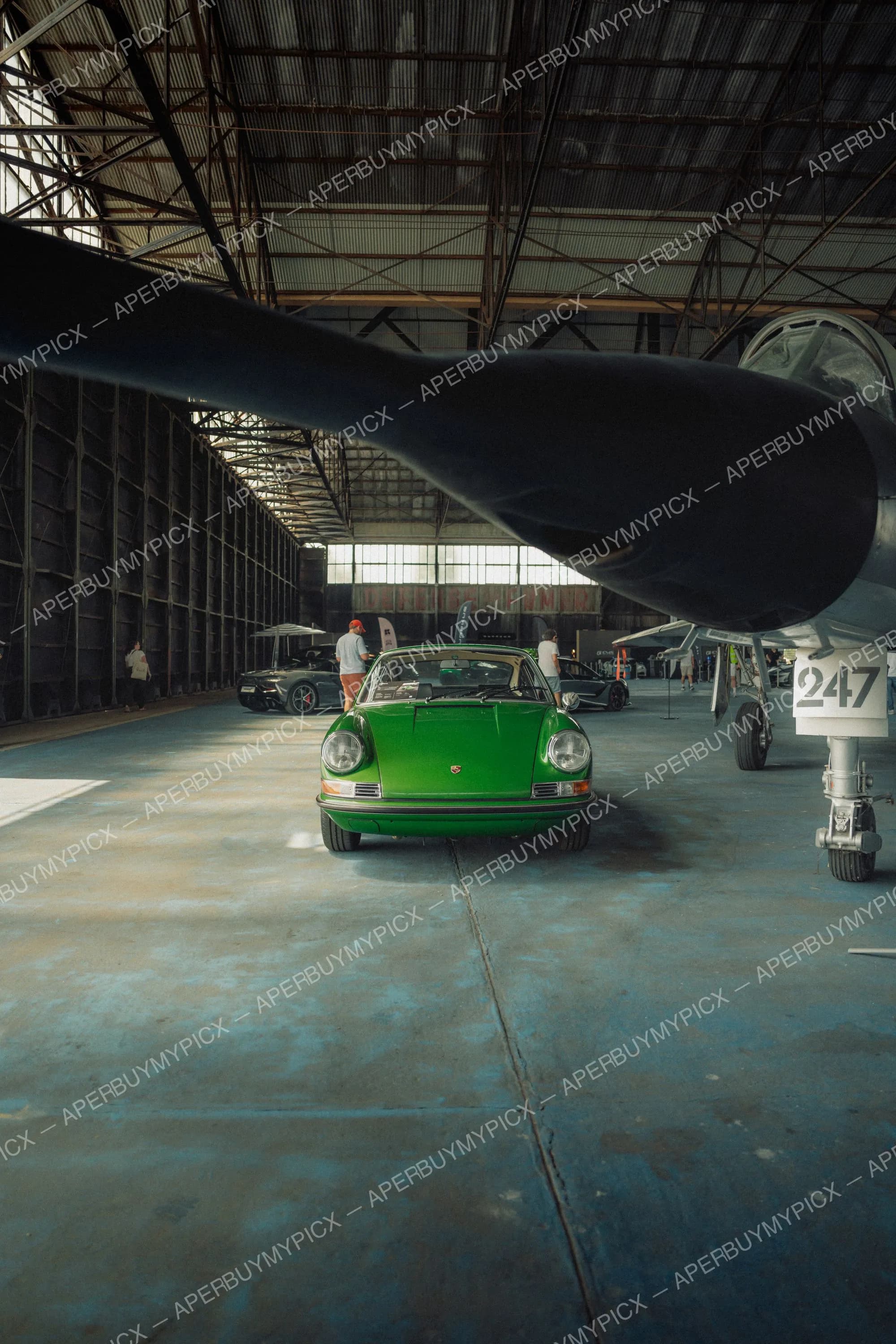 Porsche 911 vintage verte exposée dans un hangar d'aviation sous l'aile et l'hélice d'un avion militaire numéroté 247, avec d'autres voitures de sport et des visiteurs en arrière-plan