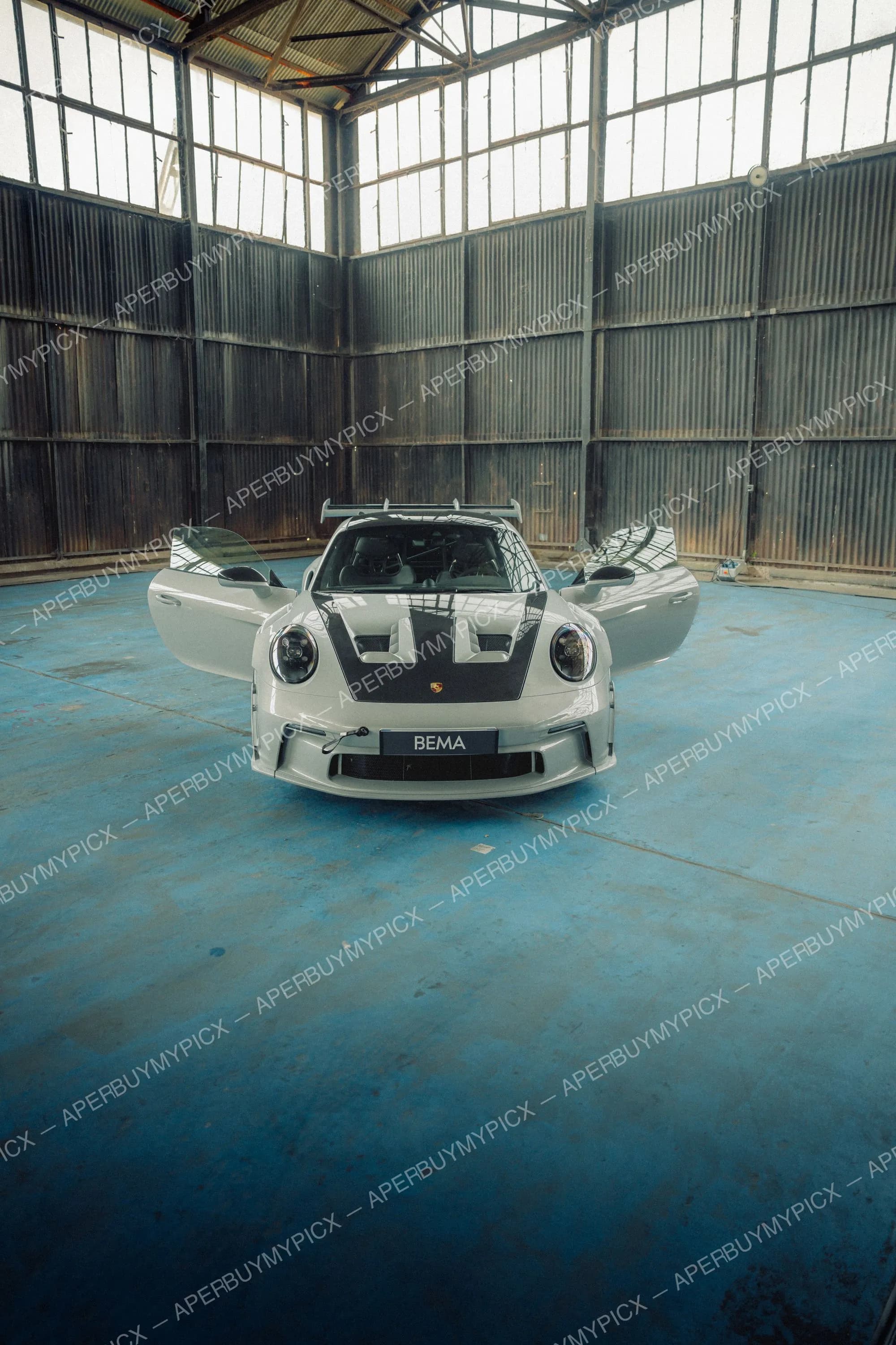 Porsche 911 GT3 RS grise vue de face avec capot en carbone et portes ouvertes, aileron arrière, garée sur sol bleu dans un hangar industriel aux grandes verrières.