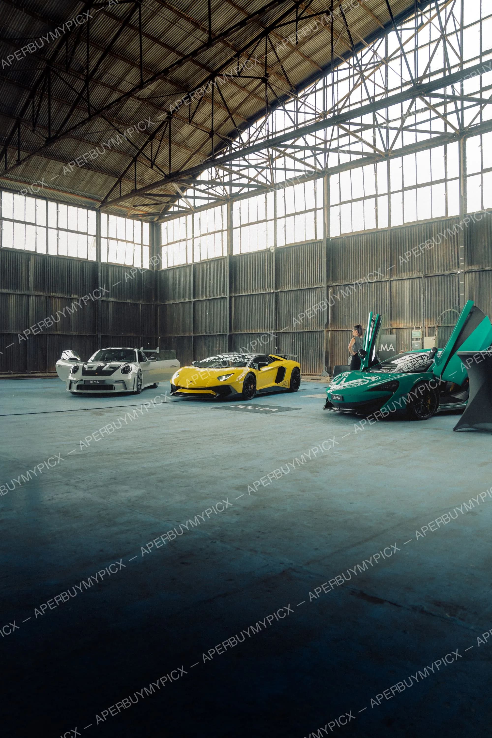 Trois supercars - une Porsche 911 blanche, une Lamborghini Aventador jaune et une McLaren verte aux portières ouvertes - dans un vaste hangar industriel avec verrière, une femme debout à côté de la voiture verte.