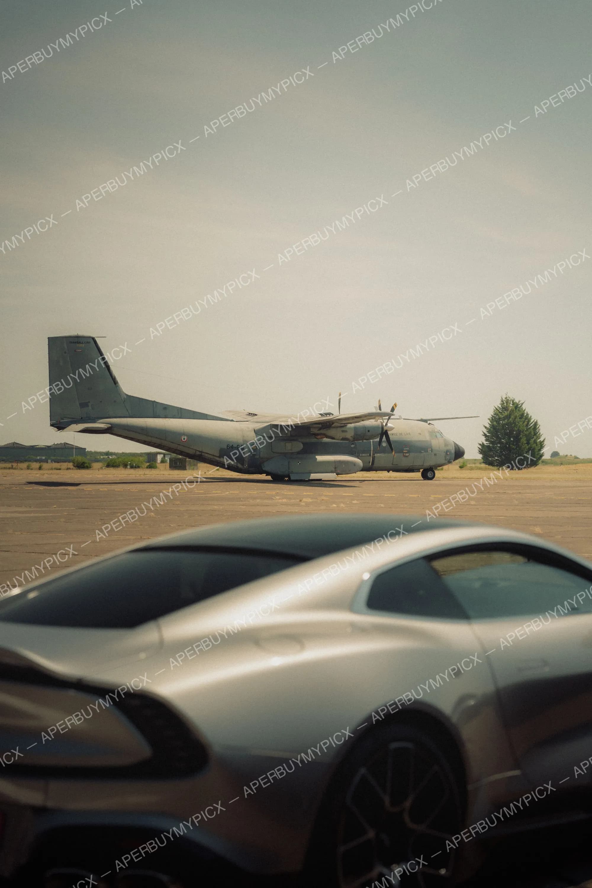 Une supercar grise au premier plan sur un tarmac avec un avion militaire Transall C160 de l'Armée de l'Air en arrière-plan sous un ciel voilé