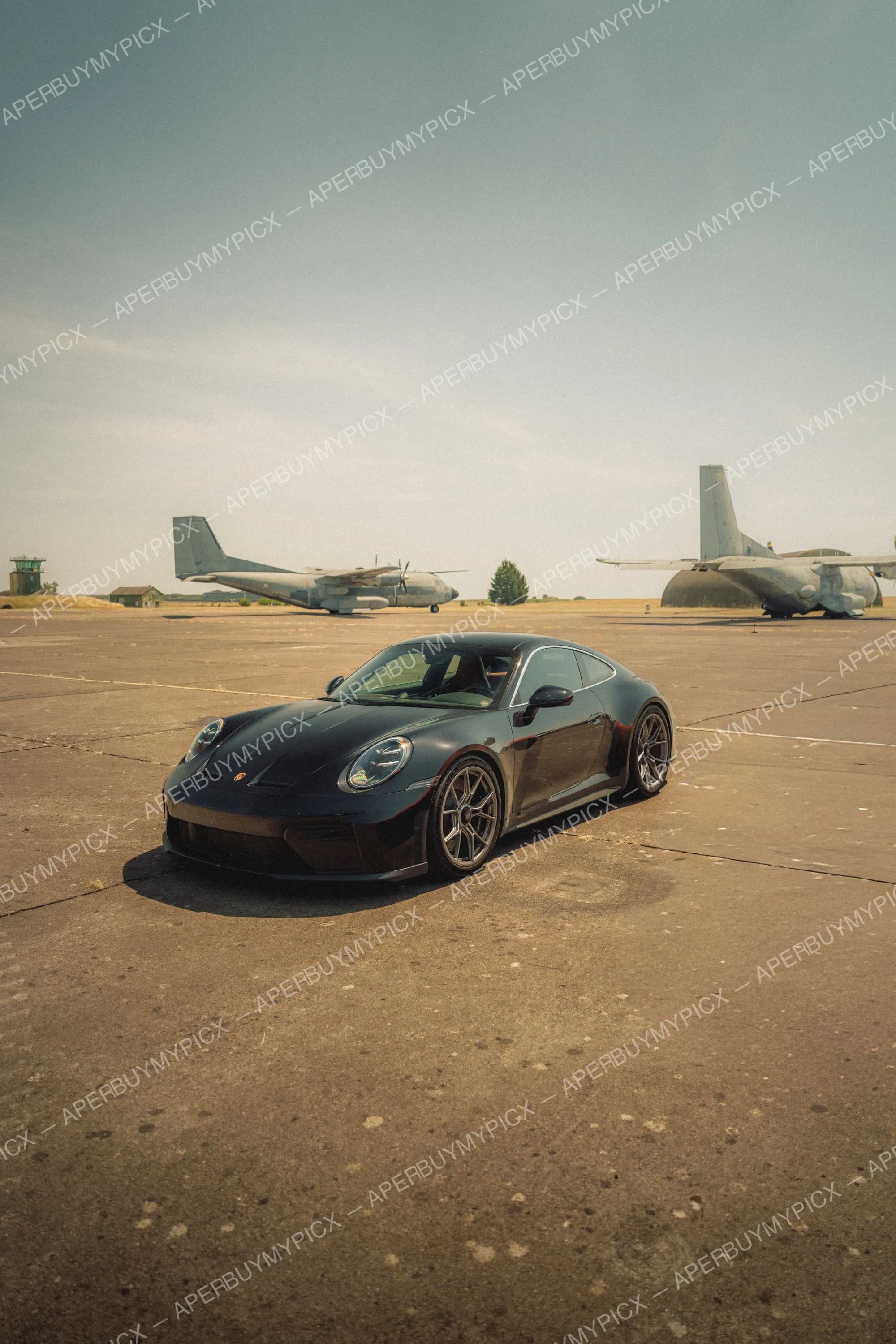 Porsche 911 GT3 noire garée sur le tarmac d'une base aérienne, avec deux avions de transport militaire à l'arrière-plan sous un ciel dégagé.