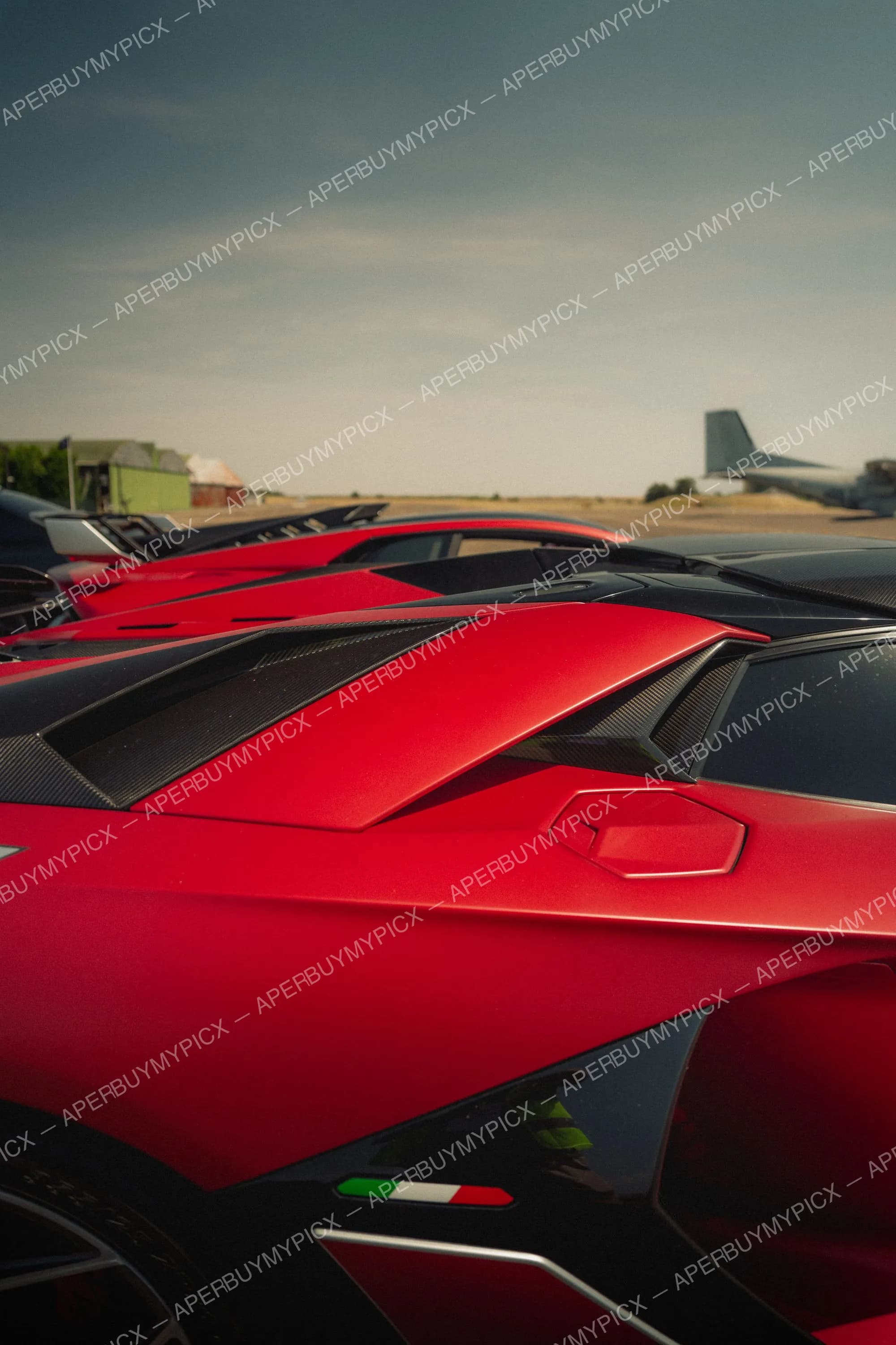 Gros plan sur le capot arrière d'une Lamborghini rouge aux inserts en fibre de carbone, alignée avec d'autres supercars sur une piste d'aérodrome, drapeau italien visible et avion en arrière-plan.