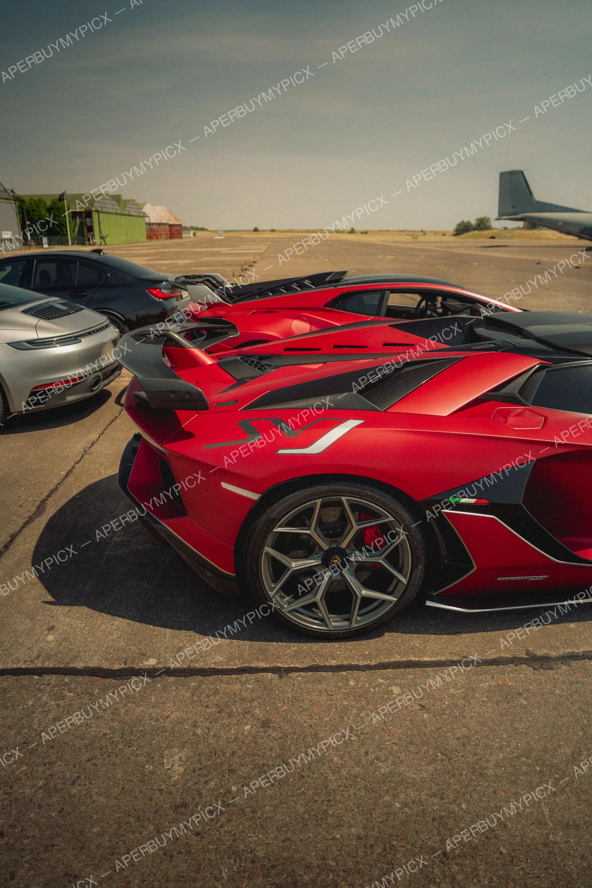 Lamborghini Aventador SVJ rouge mat avec détails en carbone et jantes grises vue de trois quarts arrière, garée sur une piste d'aviation aux côtés d'une Porsche 911 grise, d'une berline noire et d'un avion militaire sous un ciel dégagé.