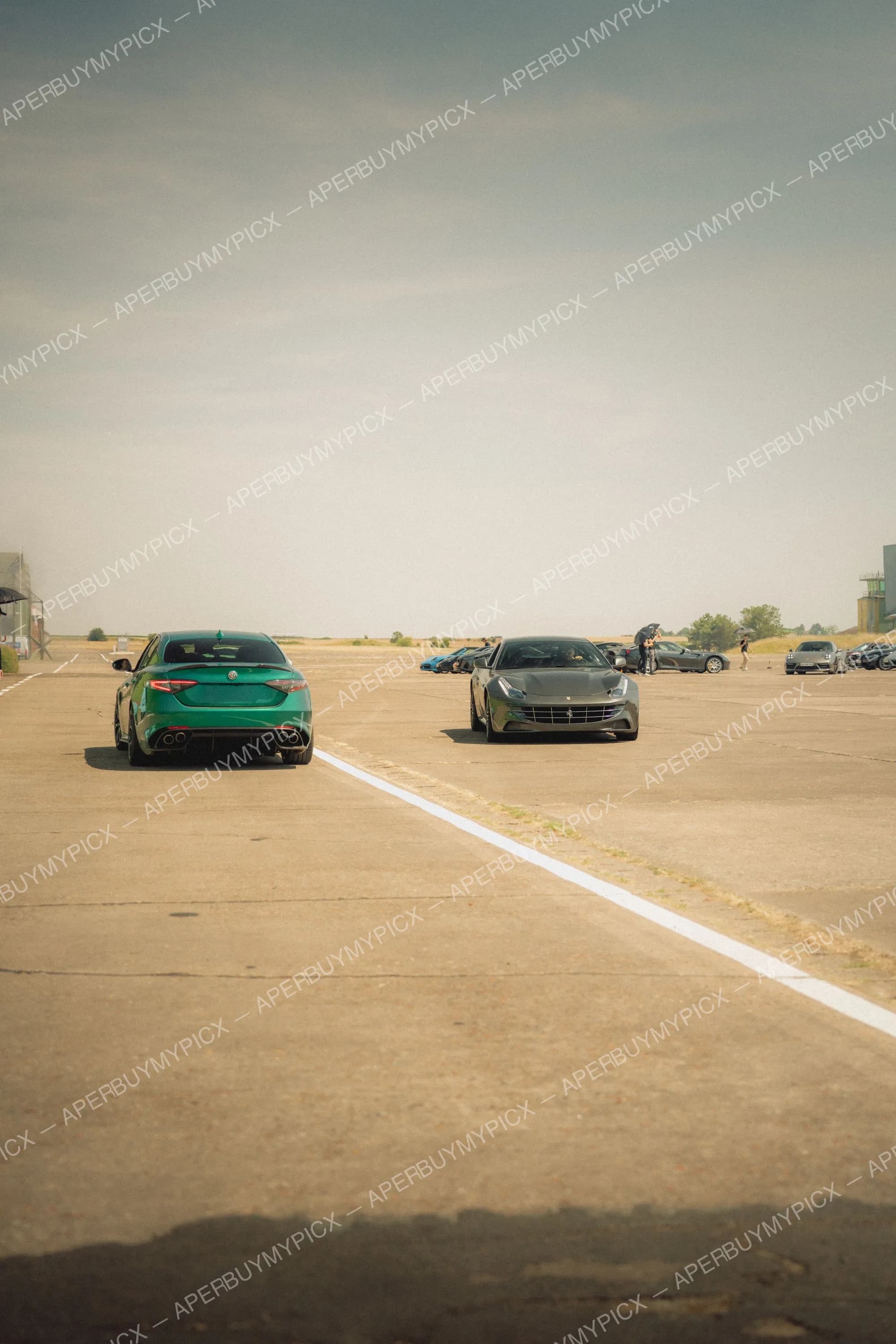 Alfa Romeo Giulia verte de dos et Ferrari FF grise de face sur une piste d'aéroport, avec d'autres voitures de sport et spectateurs au loin sous un ciel voilé.