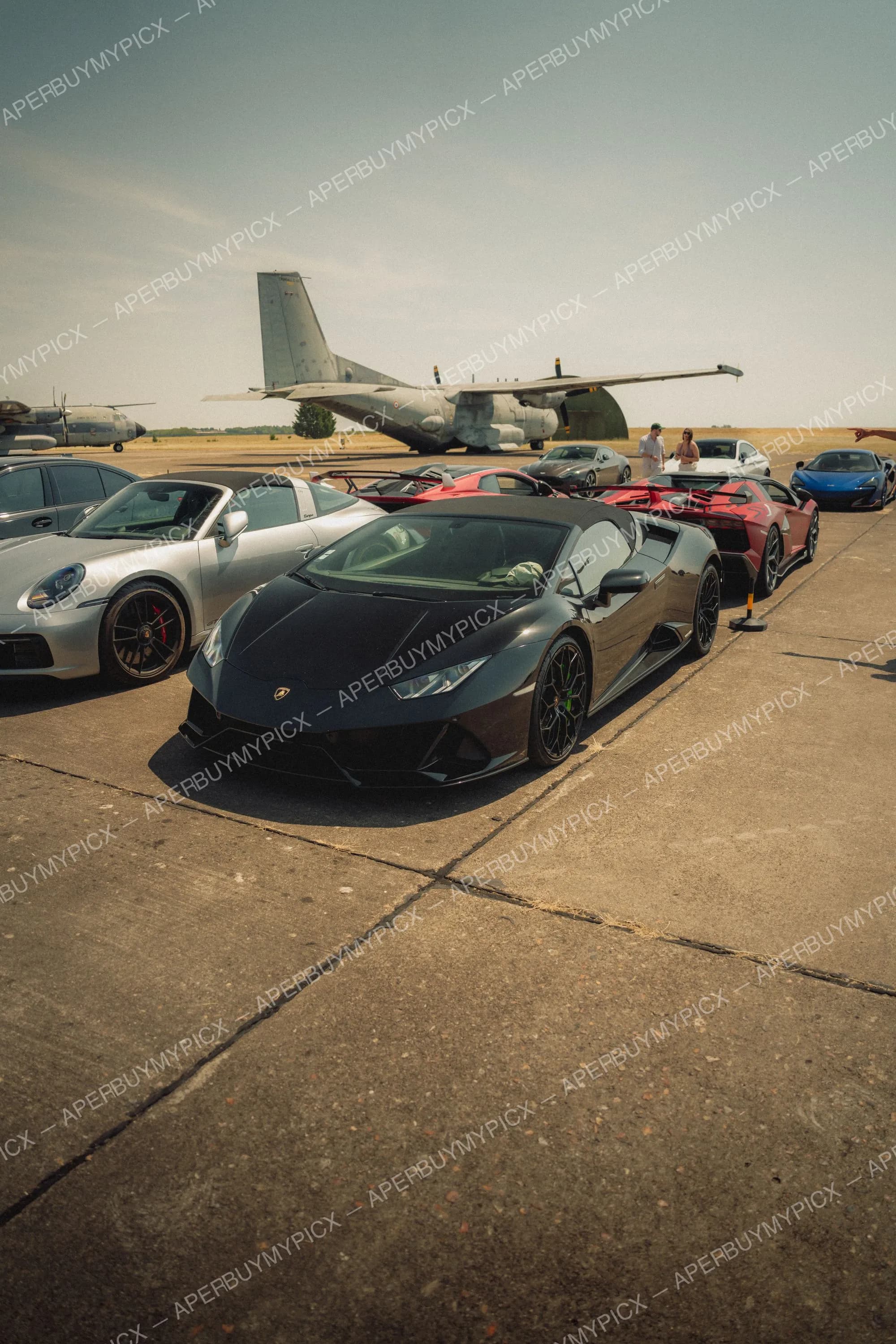 Lamborghini Huracán EVO Spyder noire garée sur le tarmac d'une base aérienne, entourée de supercars dont une Porsche 911 grise, une Aventador rouge et une McLaren bleue, avec des avions militaires en arrière-plan.