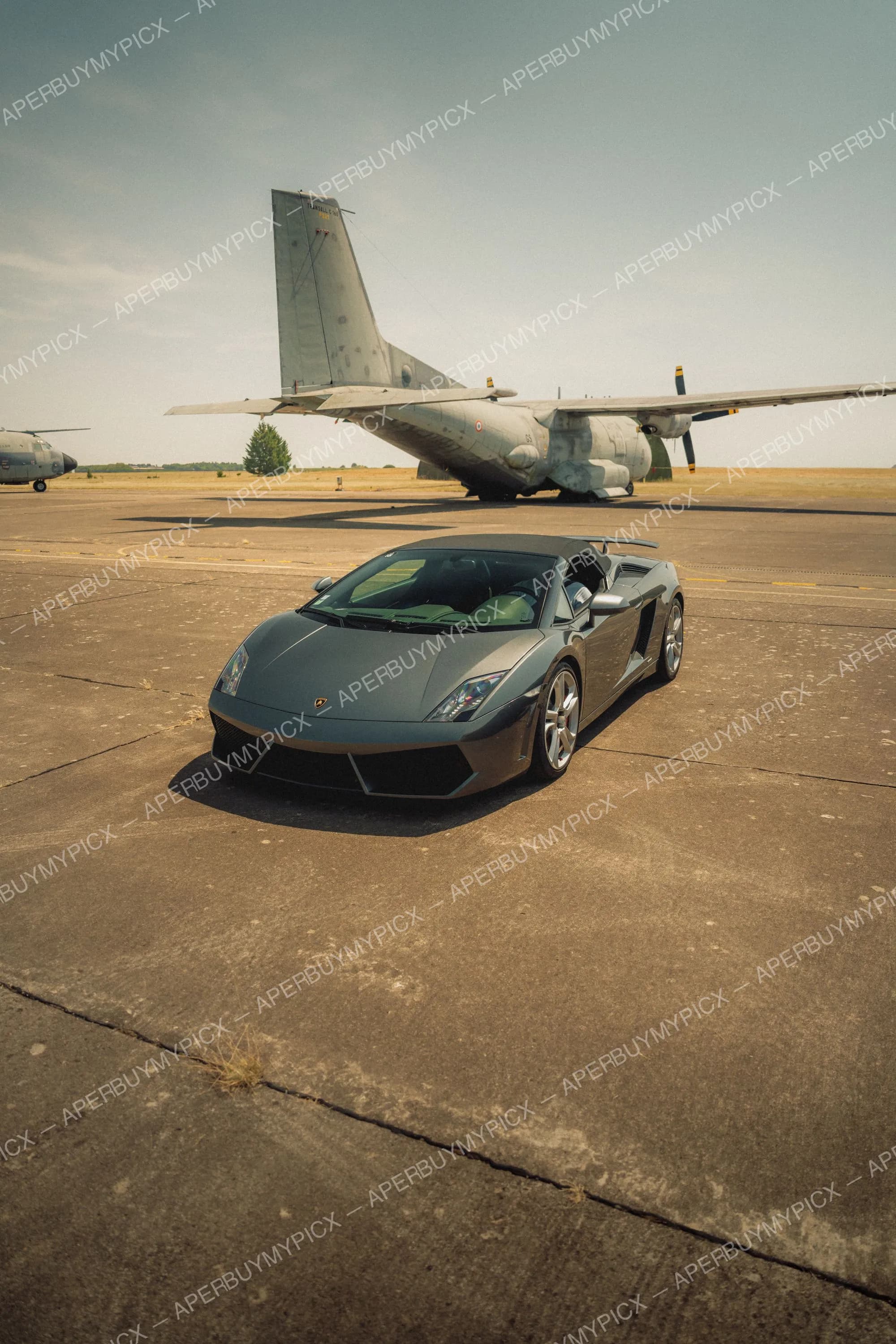 Lamborghini Gallardo grise garée sur une piste d'aérodrome militaire, avec un avion de transport Transall C-160 et un hélicoptère en arrière-plan sous un ciel clair.