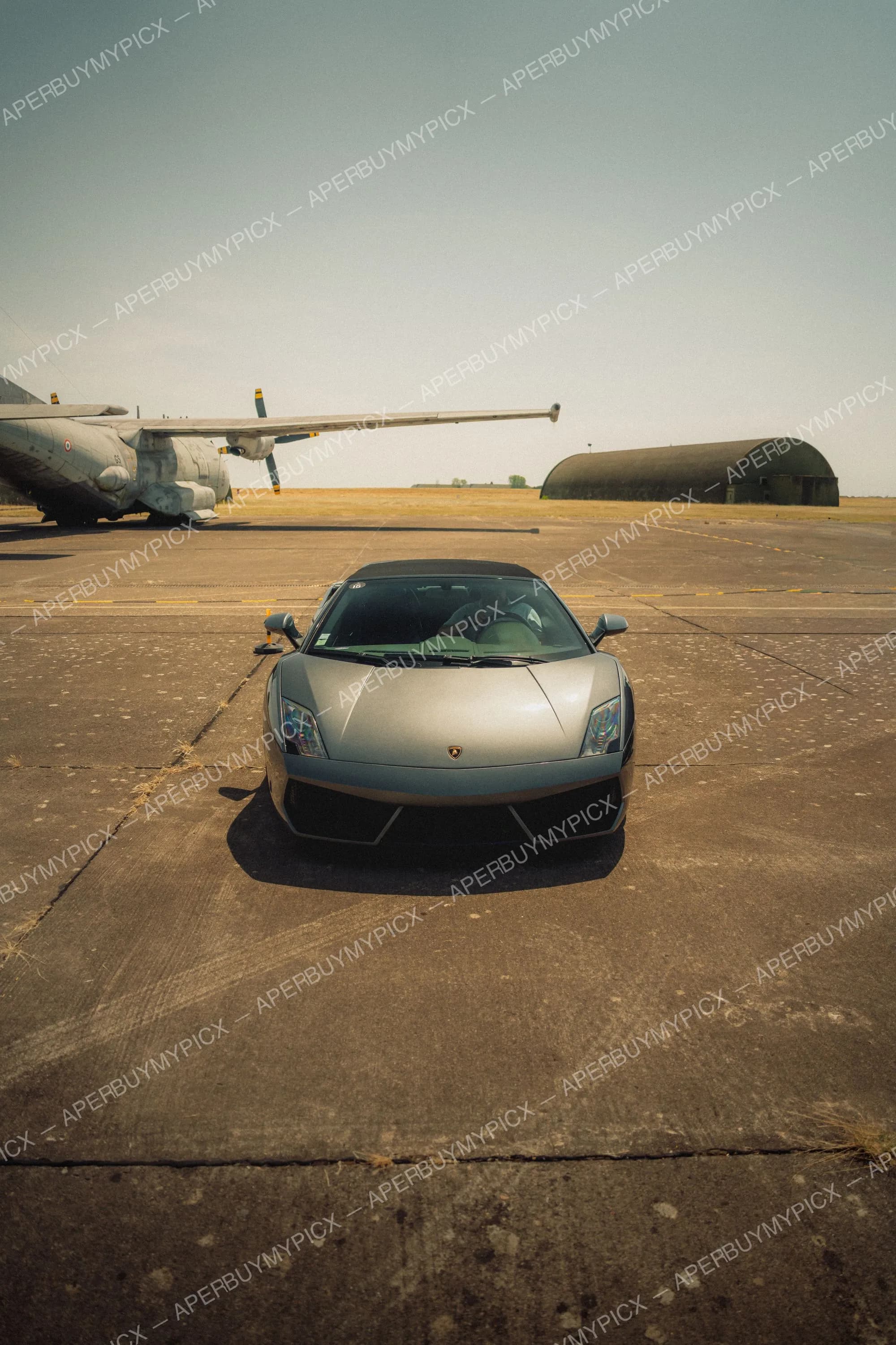 Lamborghini Gallardo grise vue de face, garée sur une piste d'aérodrome militaire avec un avion de transport à hélices et un hangar en arrière-plan sous un ciel dégagé.