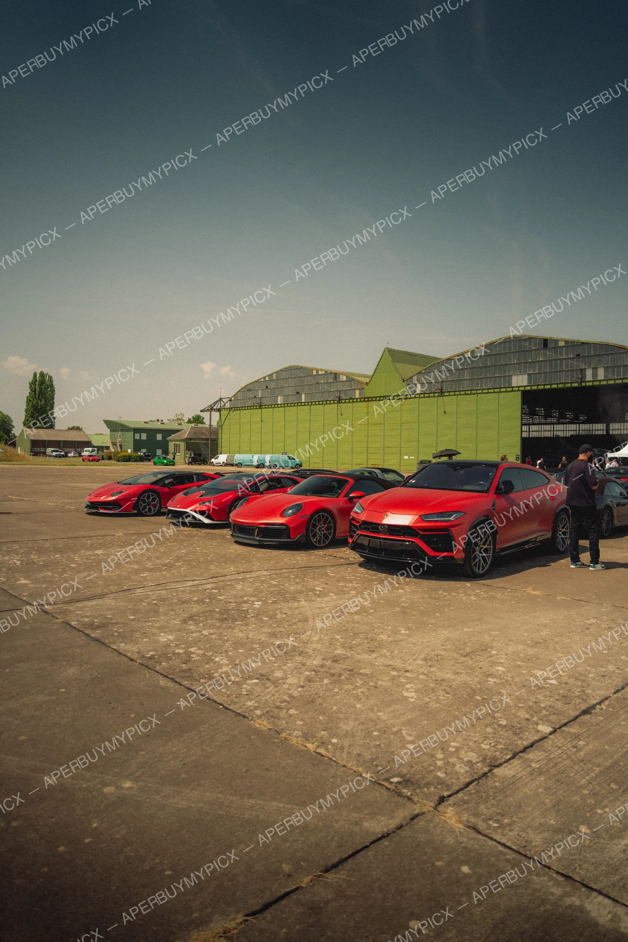 Rangée de supercars rouges dont une Lamborghini Aventador, une Huracán, une Porsche 911 Turbo et un Lamborghini Urus, garées sur le tarmac devant un grand hangar vert par temps ensoleillé.