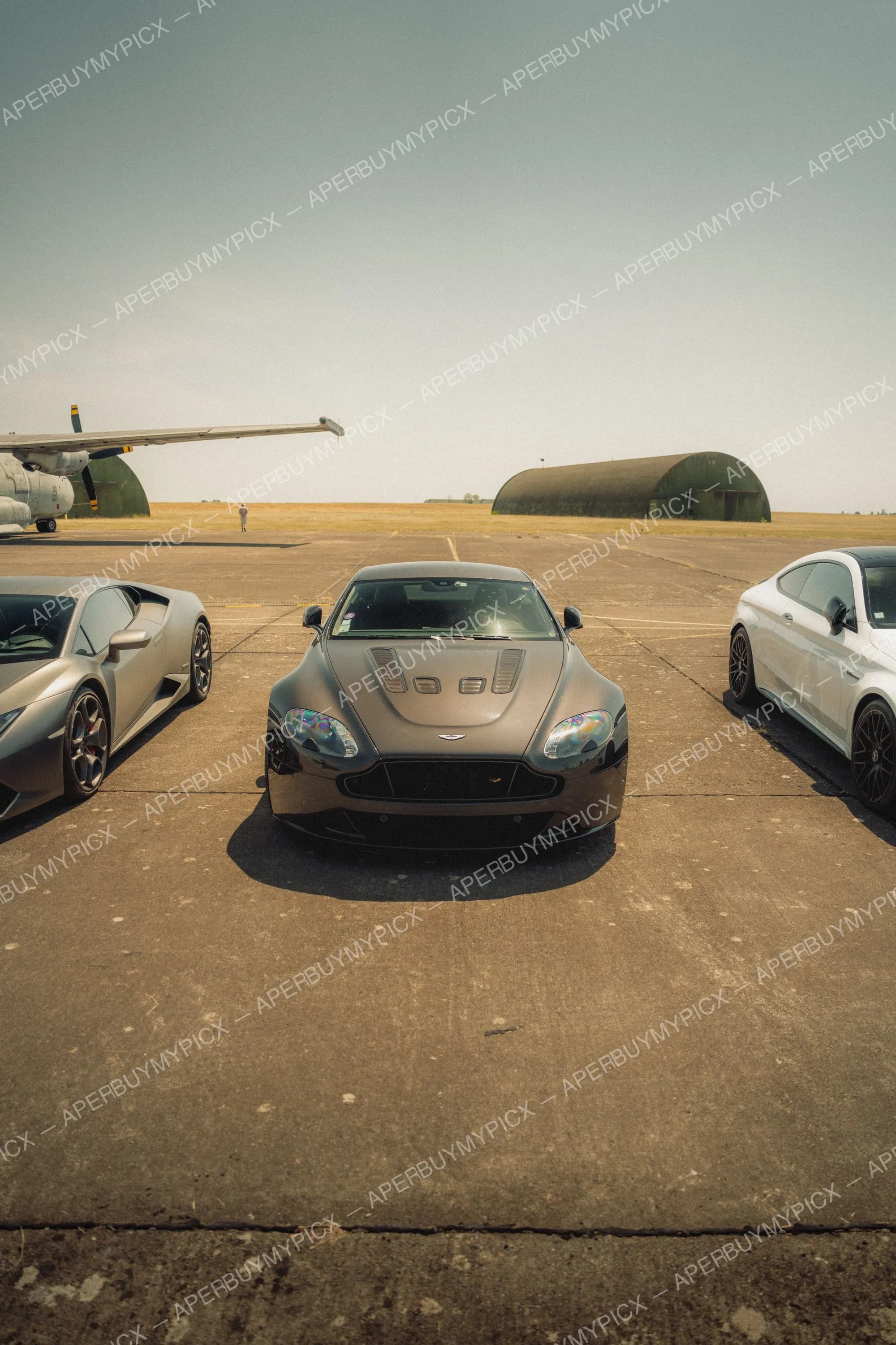 Aston Martin V12 Vantage S grise vue de face sur une piste d'aéroport, encadrée par une Lamborghini Huracán argentée et une Mercedes AMG blanche, avec avions et hangars militaires en arrière-plan sous un ciel dégagé.