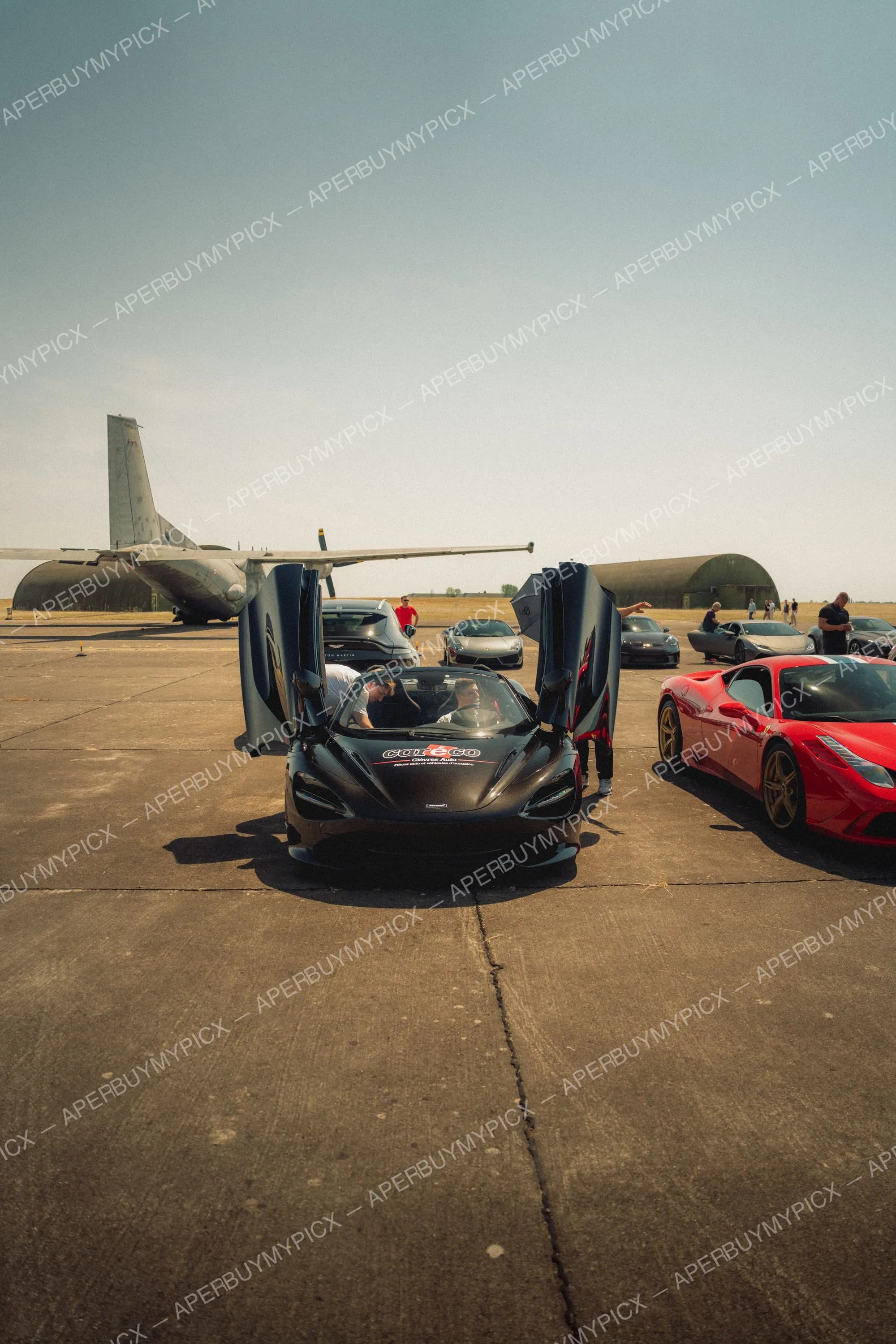 McLaren 720S noire aux portes papillon ouvertes garée sur une piste d'aéroport, à côté d'une Ferrari 458 Speciale rouge, avec un avion de transport militaire et des hangars en arrière-plan sous un ciel dégagé.