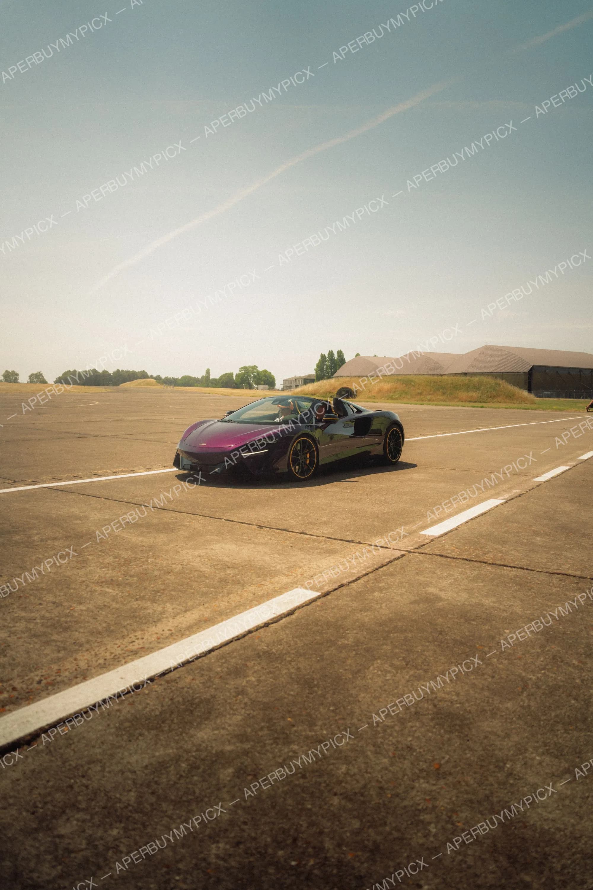 McLaren Artura Spider convertible violette dégradée avec jantes noires roulant sur une piste d'aviation en béton par temps ensoleillé, hangars à l'arrière-plan