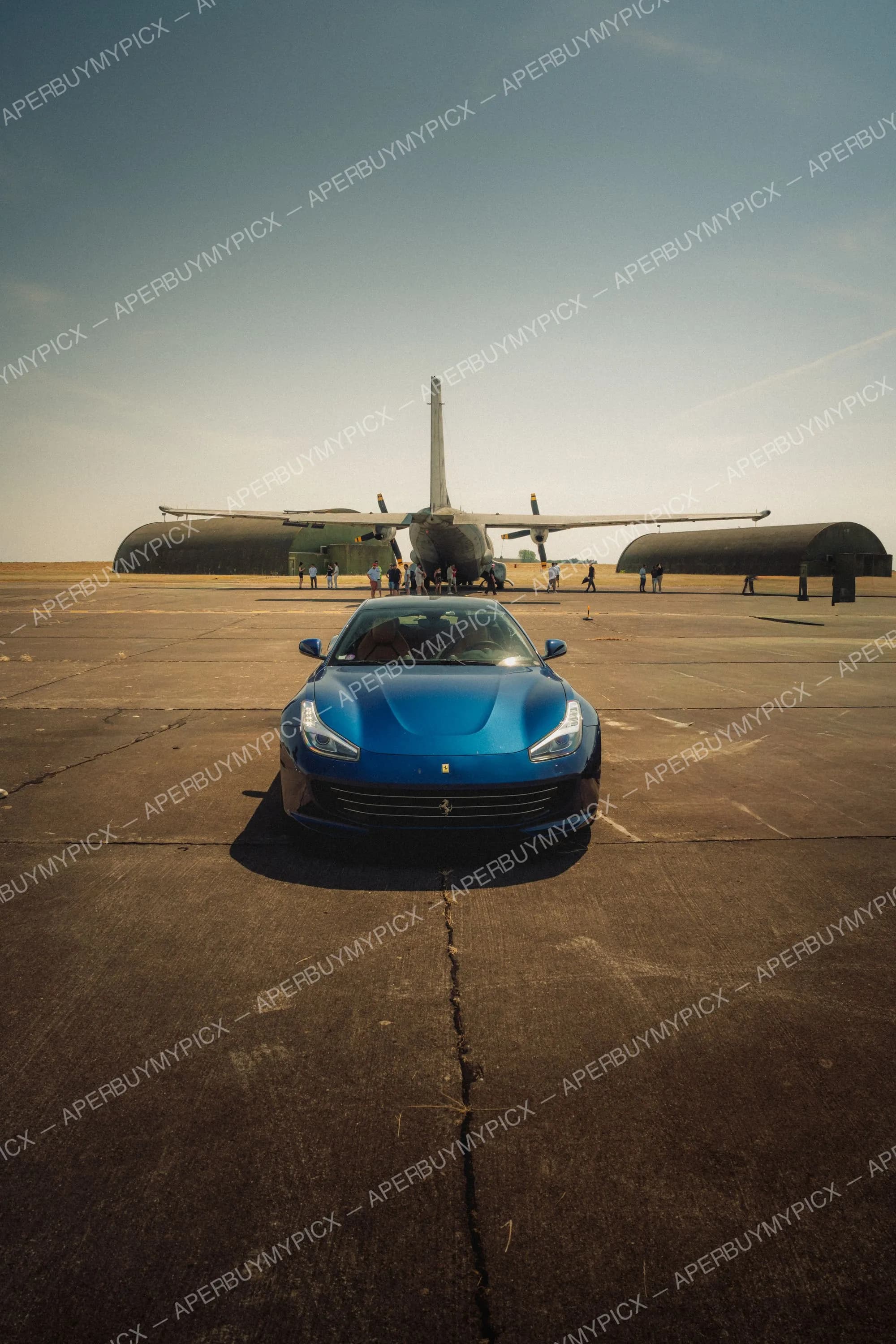 Ferrari GTC4Lusso bleue vue de face garée sur une piste d'aviation en béton, avec un avion de transport militaire à hélices et des hangars en arrière-plan sous un ciel dégagé.
