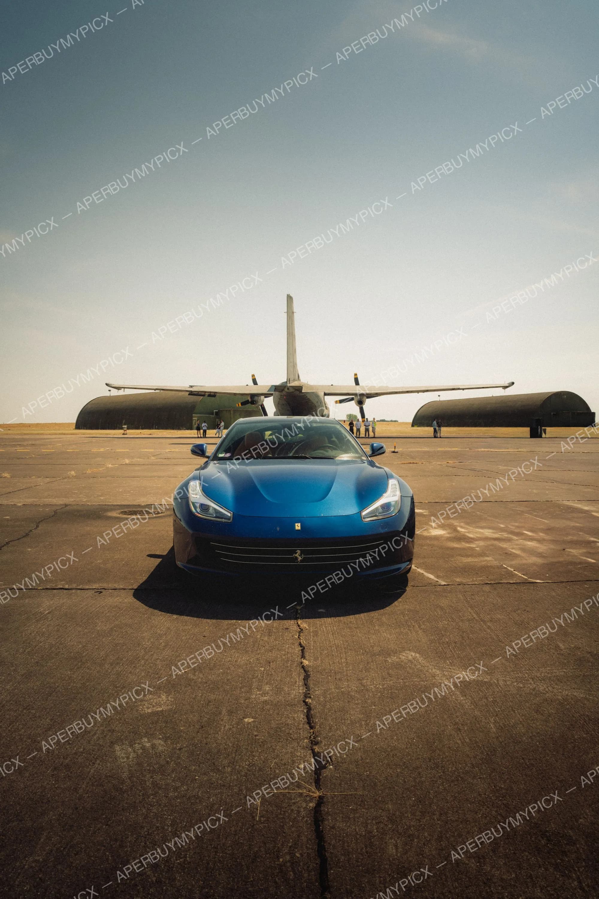 Ferrari GTC4Lusso bleue vue de face garée sur une piste d'aéroport en béton, avec un avion militaire à hélices et des hangars en arrière-plan sous un ciel dégagé.