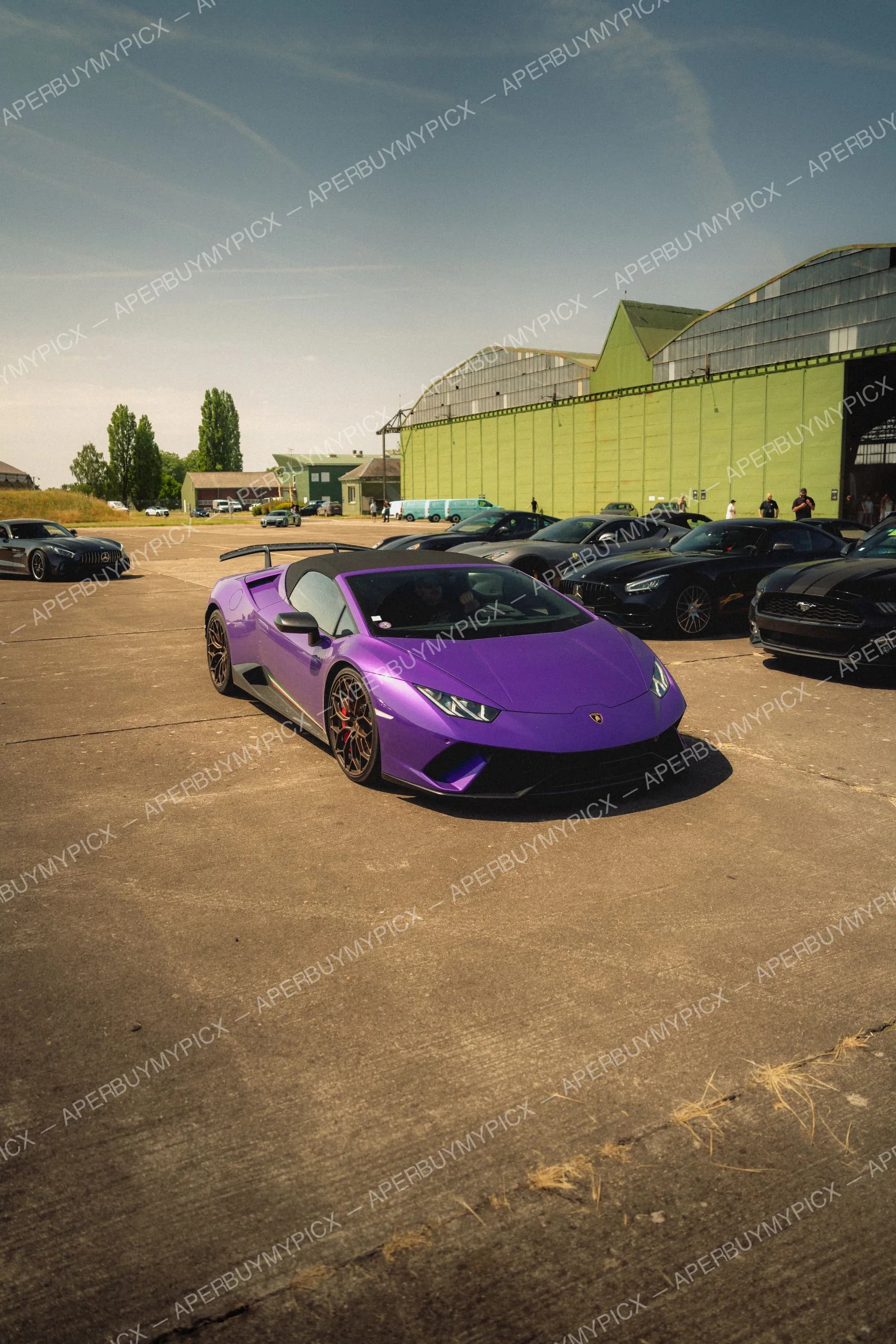 Lamborghini Huracán Performante violette avec jantes dorées garée sur un tarmac devant un hangar vert, entourée de supercars noires dont une Mercedes-AMG GT et une Ford Mustang.