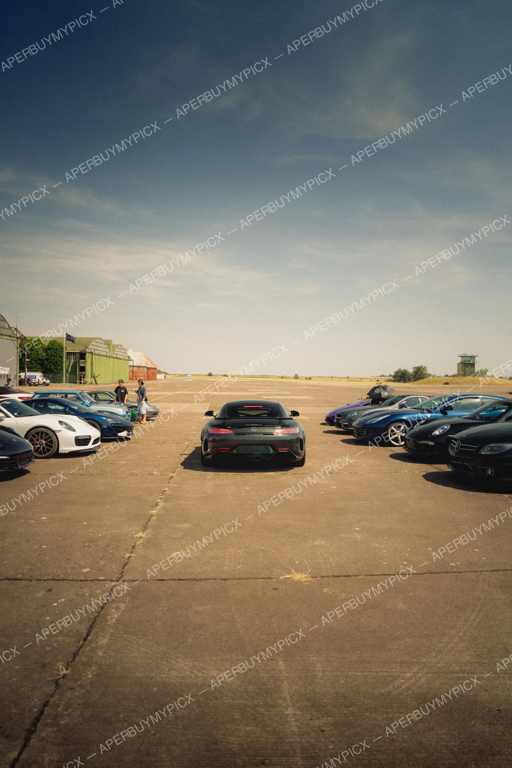 Mercedes-AMG GT noire vue de l'arrière au centre d'une piste d'aérodrome, entourée de supercars alignées dont des Porsche 911, une Ferrari et une McLaren, avec des hangars verts et un ciel bleu en arrière-plan.
