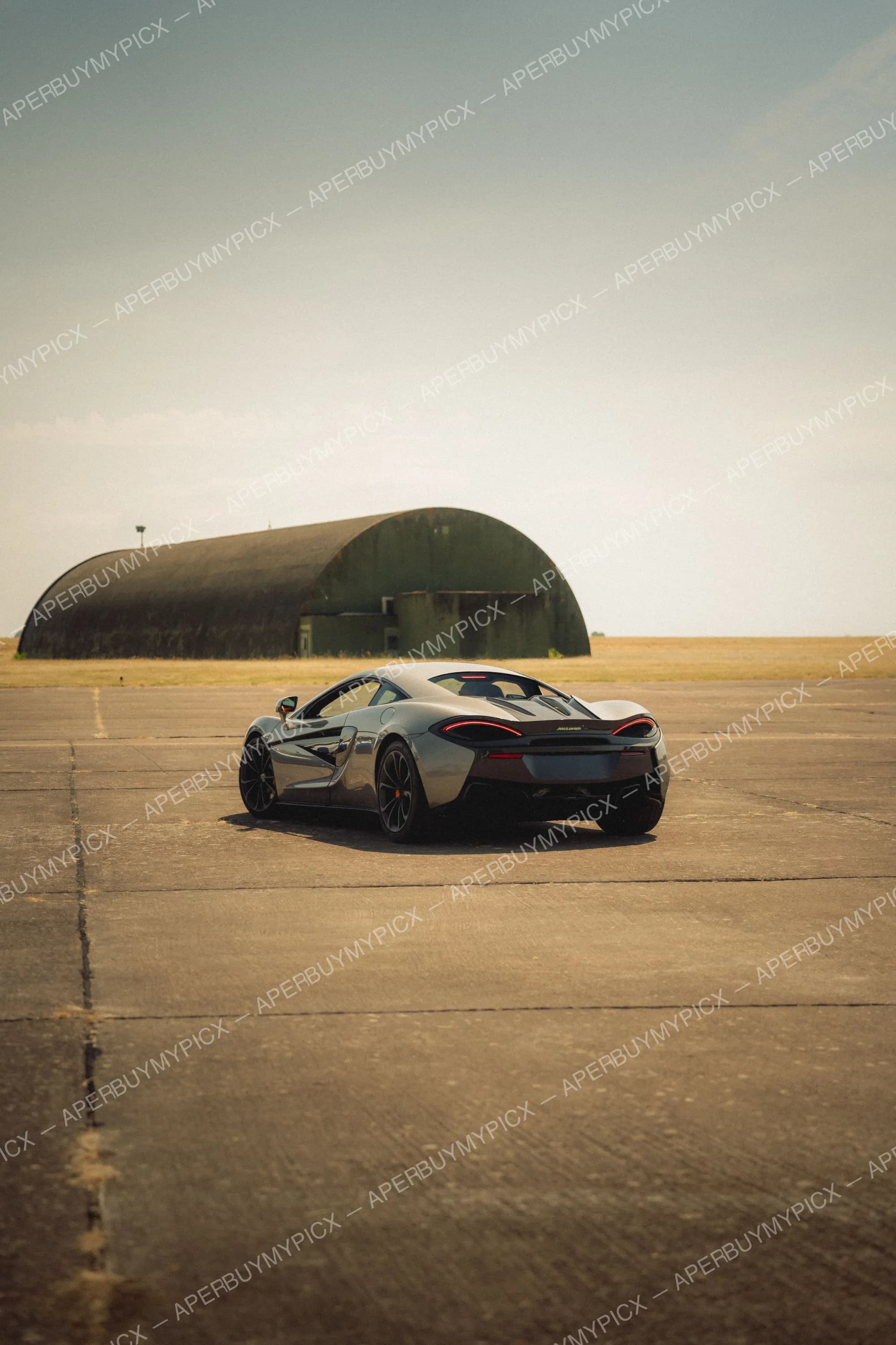 McLaren 570S grise vue de trois quarts arrière, garée sur une piste en béton d'un ancien aérodrome, avec un hangar voûté vert et un ciel clair en arrière-plan.